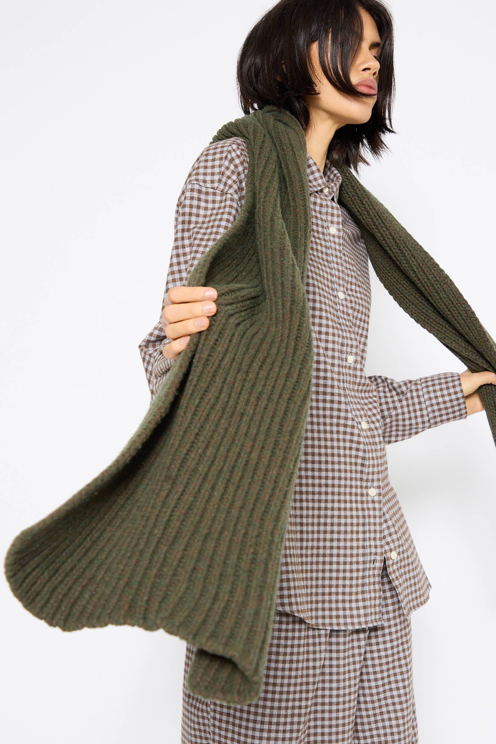 The person, dressed in a brown checkered outfit, presents the Mea Scarf in Foret by Baserange—a long, chunky knit scarf crafted from a recycled wool blend in dark green.