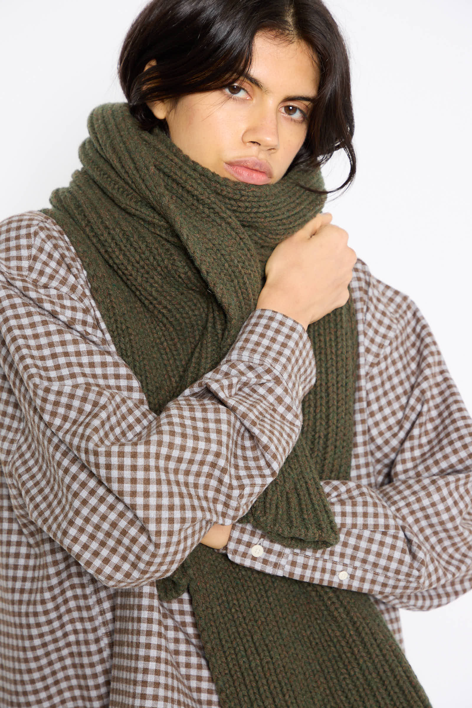 A person wearing a plaid shirt and the Baserange Mea Scarf in Foret, made from a recycled wool blend, looks at the camera with arms crossed.