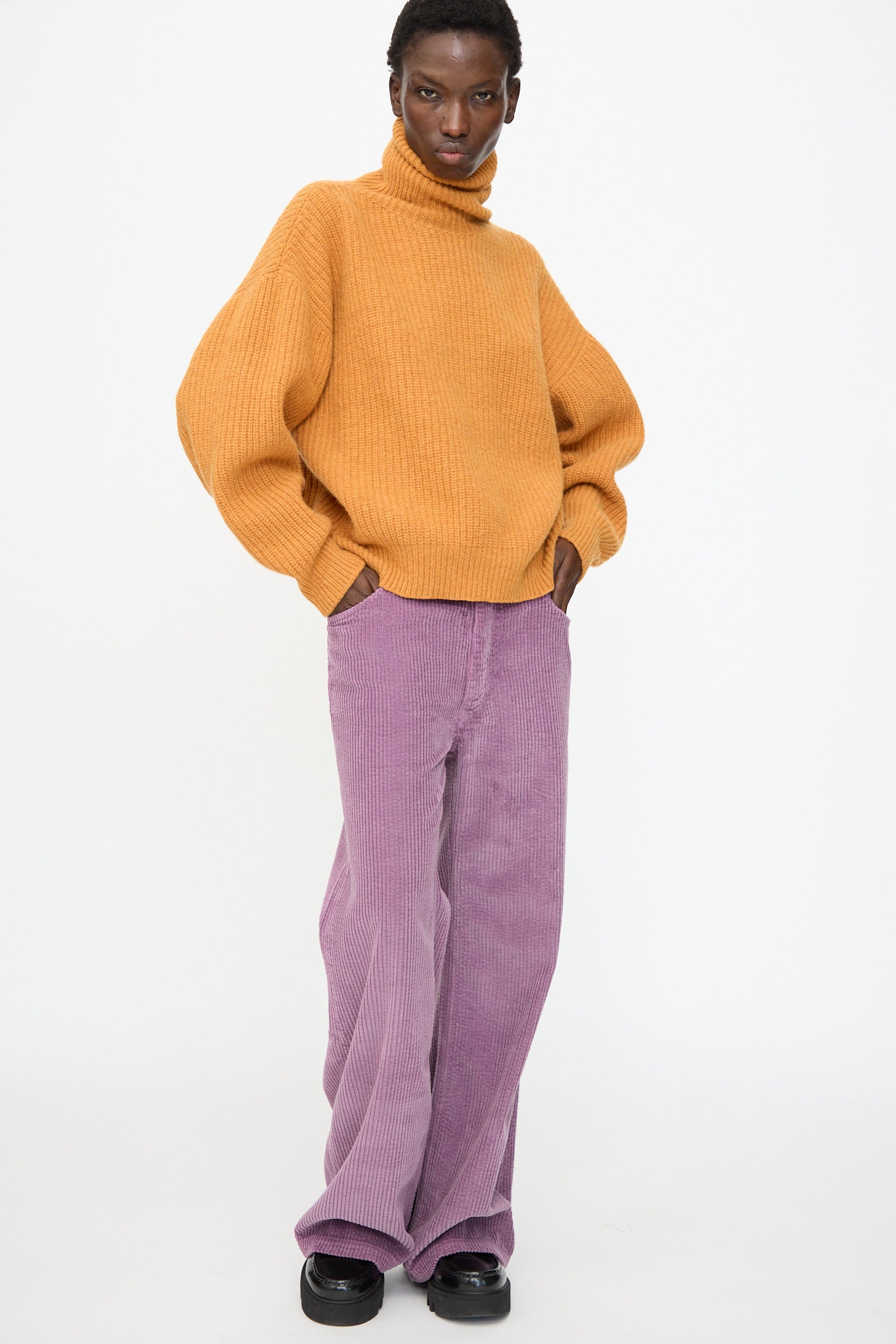 A person wearing the Baserange Mea Turtleneck in Clair Orange, loose lilac corduroy pants, and black shoes stands against a plain white background.