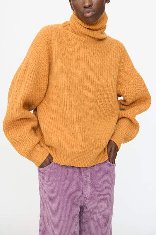 A person wears the Baserange Mea Turtleneck in Clair Orange with light purple corduroy pants, standing with hands in pockets against a plain background.