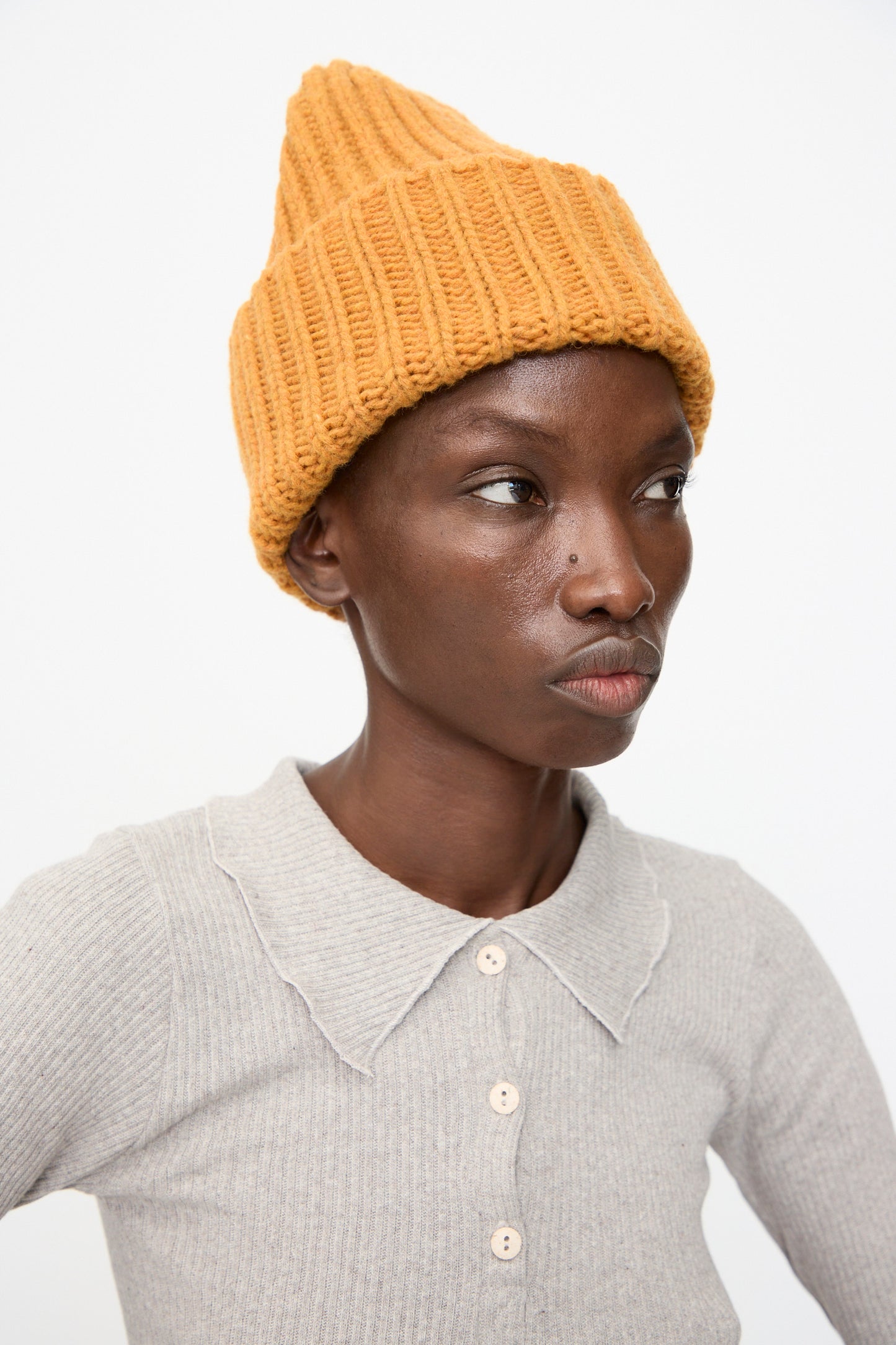A person wears the Baserange Mea Beanie in Clair Orange with a light gray, scallop-collared, buttoned shirt, looking slightly to the side against a plain background.