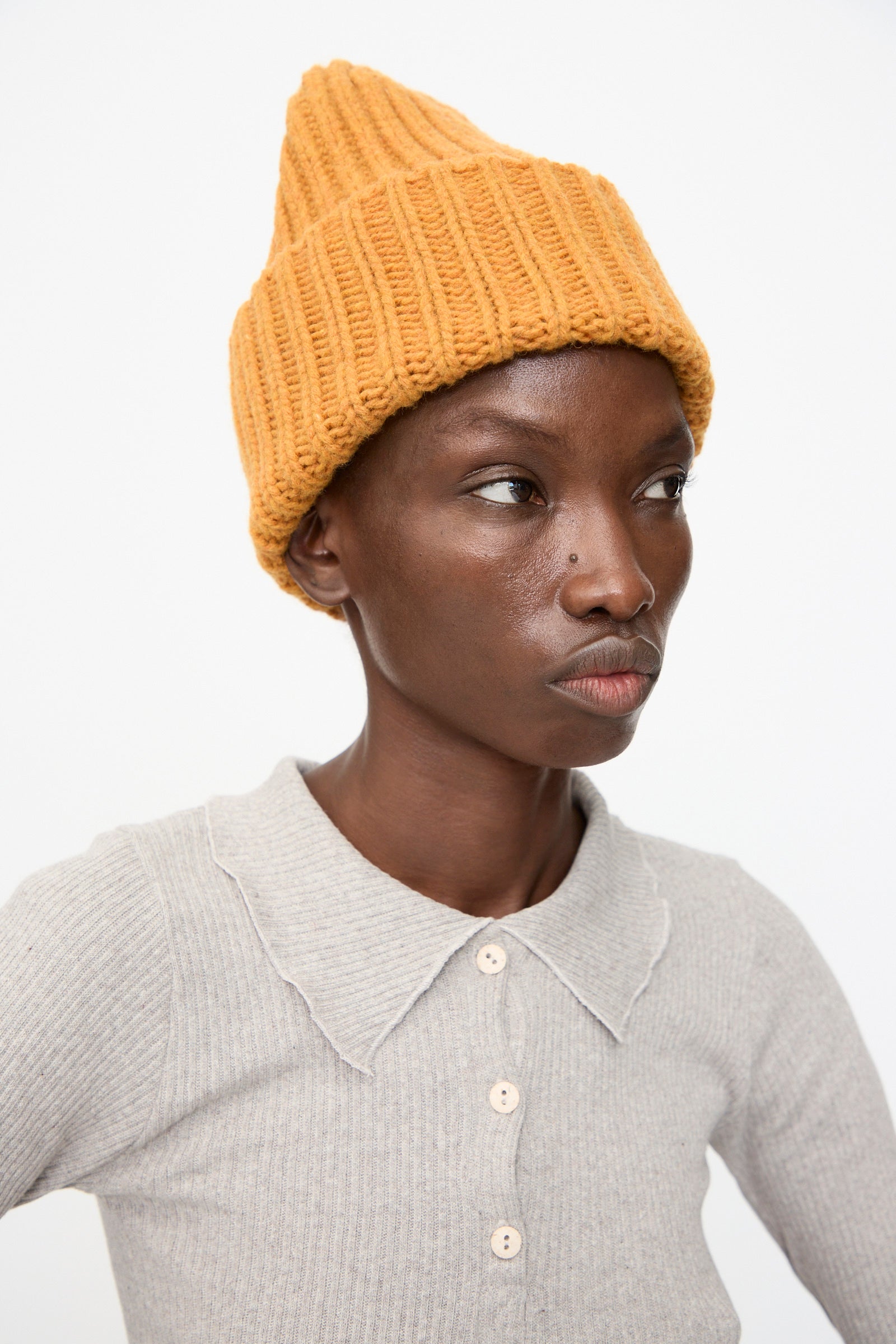 A person wears the Baserange Mea Beanie in Clair Orange with a light gray, scallop-collared, buttoned shirt, looking slightly to the side against a plain background.