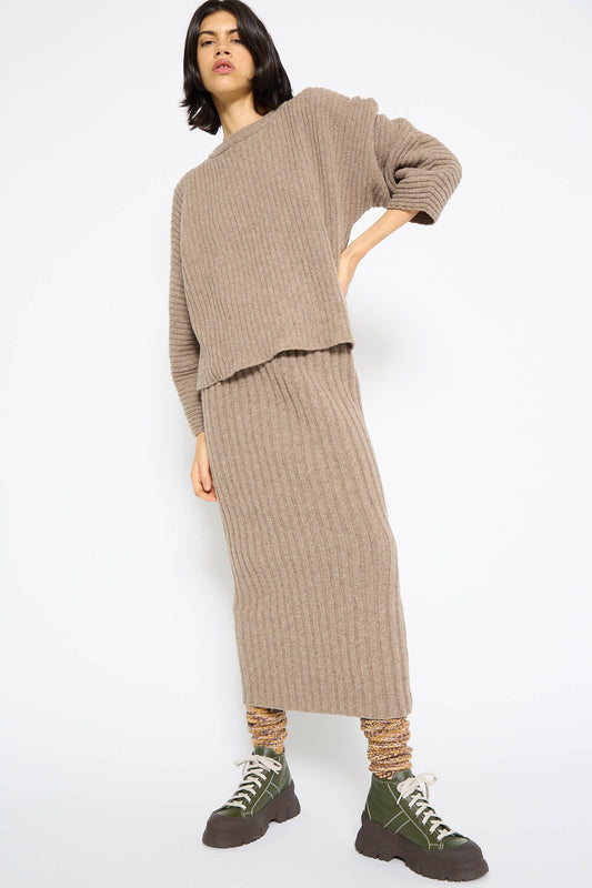 A person standing against a white background, wearing a beige merino wool ribbed knit sweater paired with the Merino Wool Kai Skirt in Alassio from Baserange, with an elasticated waist, accessorized with green boots and patterned socks.