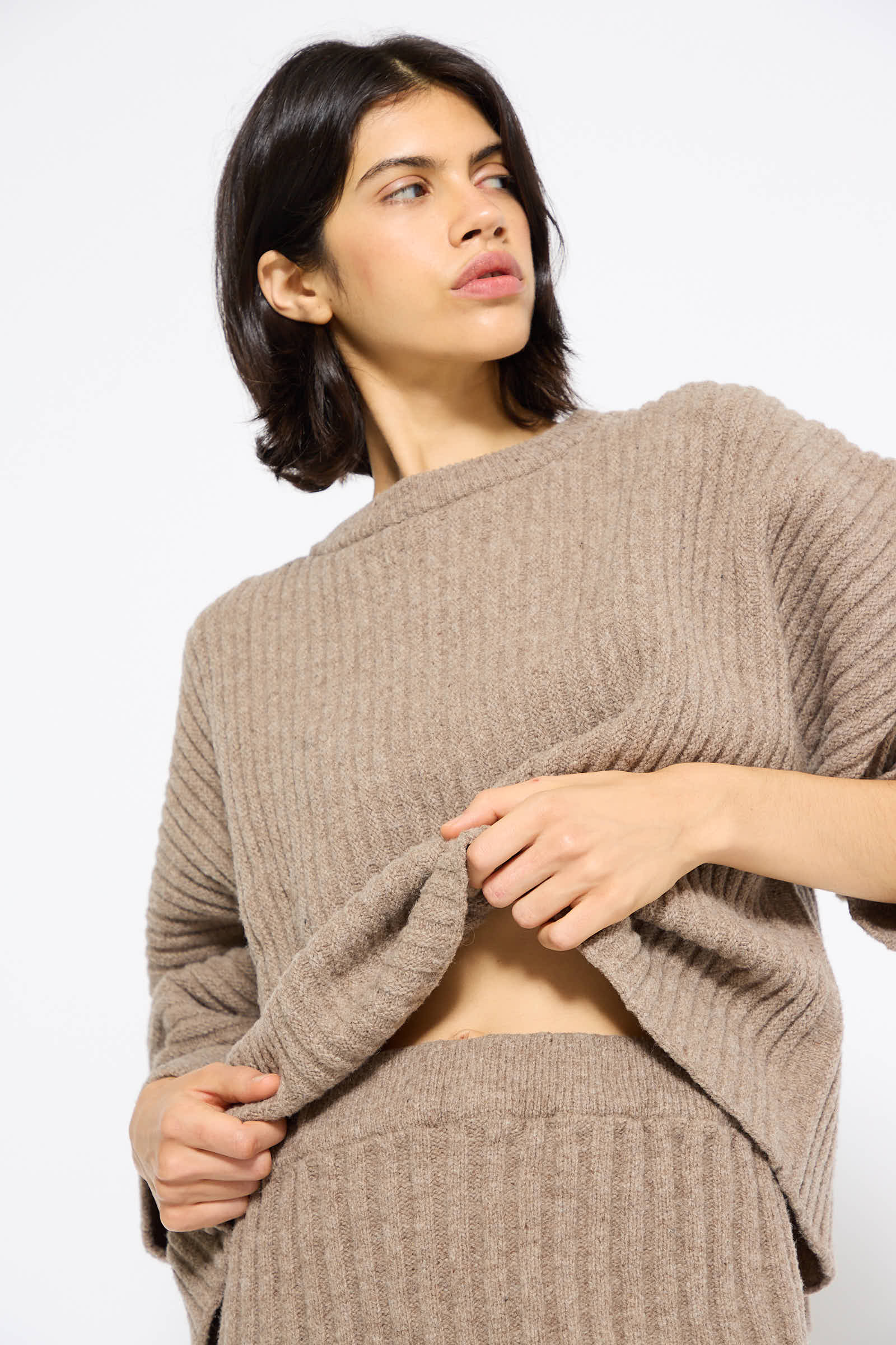 A person with short dark hair stands against a plain background, wearing a ribbed, beige knit top and the Merino Wool Kai Skirt in Alassio from Baserange, pulling up the hem of the top slightly to reveal a hint of midriff.