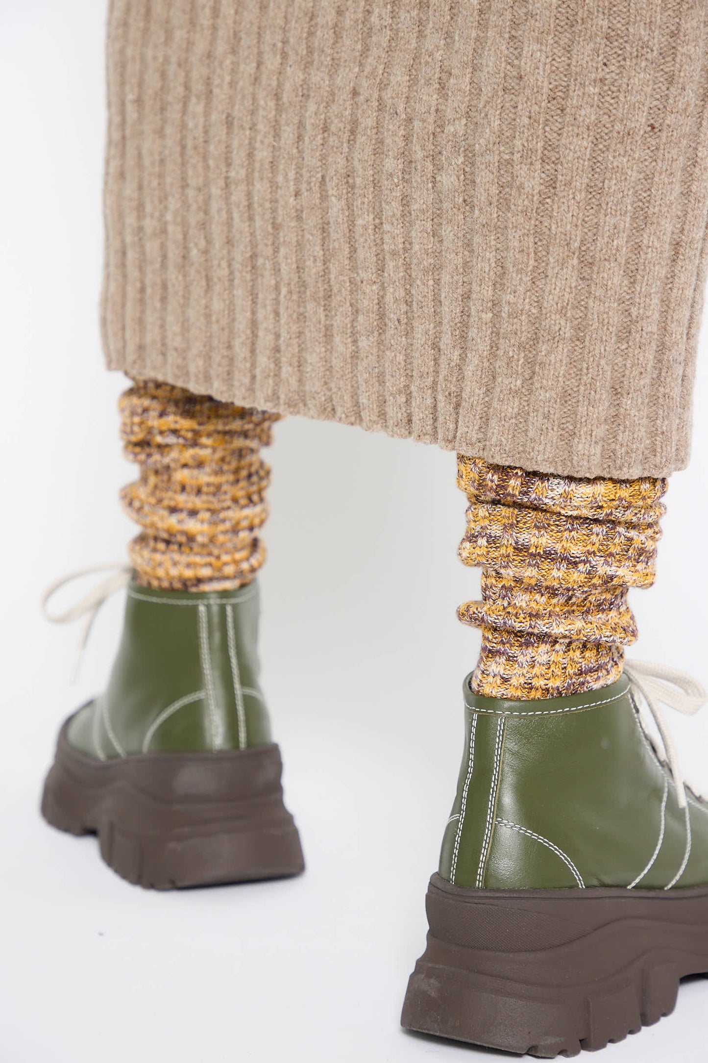 A close-up of a person wearing green lace-up chunky shoes, golden-yellow speckled socks, and the Merino Wool Kai Skirt in Alassio by Baserange, featuring a beige ribbed texture and an elasticated waist.