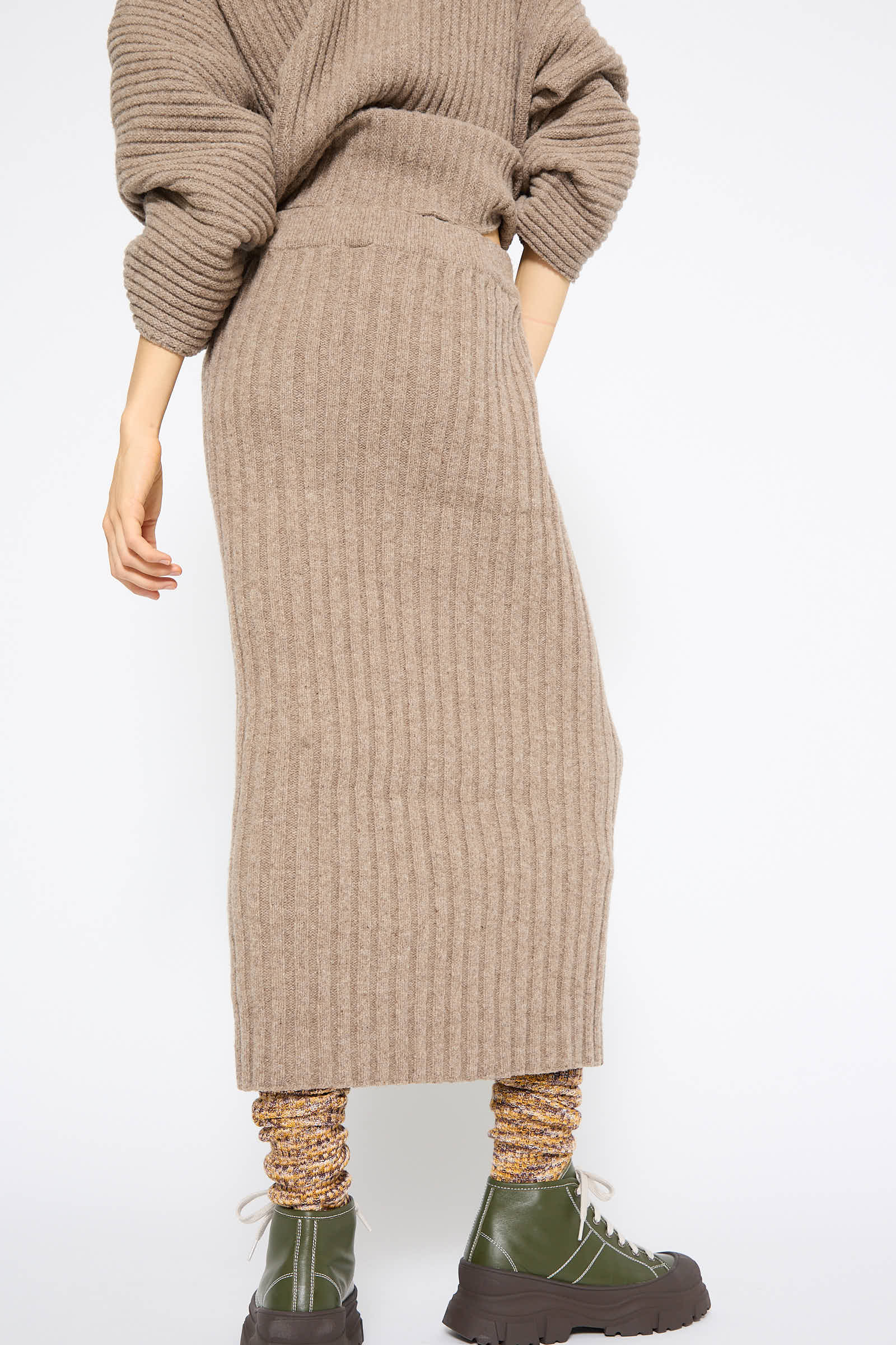 A person wearing the Merino Wool Kai Skirt in Alassio, a long, ribbed, beige merino wool knit skirt by Baserange with an elasticated waist, along with a sweater, multi-colored socks, and green platform lace-up boots stands facing away from the camera.