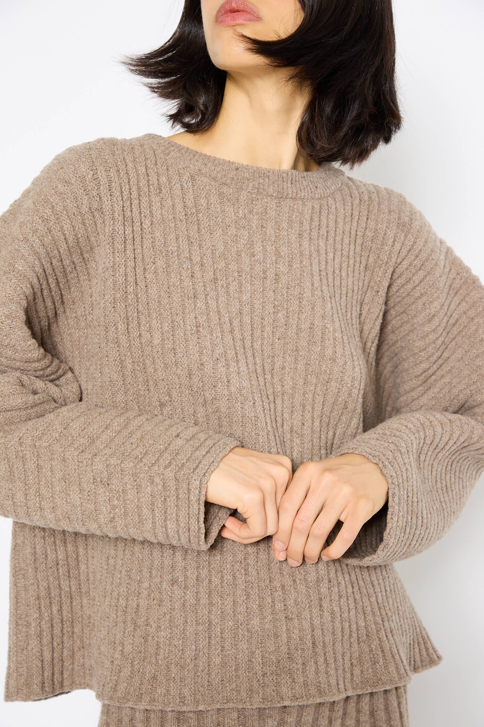 A person wearing a loose-fitting, chunky knit Baserange Merino Wool Kai Sweater in Alassio with matching pants stands against a plain white background. The person's hands are adjusting the sweater's hem, their face mostly obscured by hair.