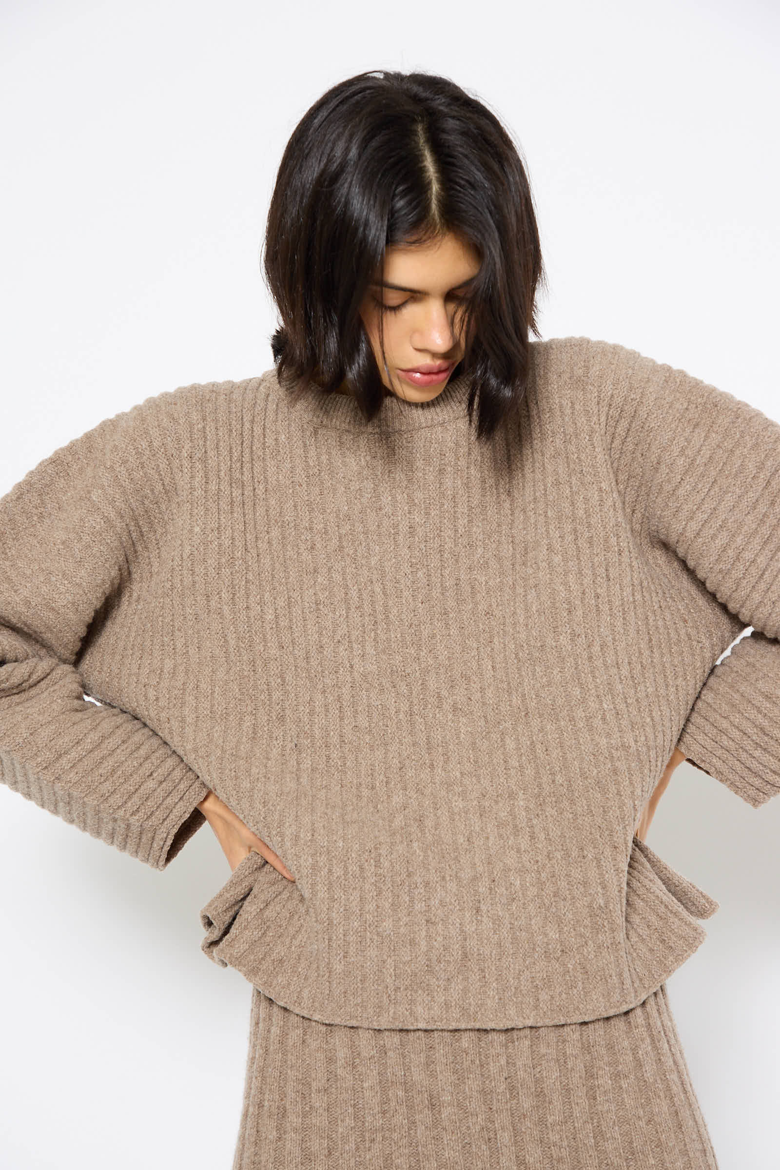 A person with medium-length dark hair wearing a beige Merino Wool Kai Sweater in Alassio by Baserange and matching skirt looks down, with hands placed on the sides.