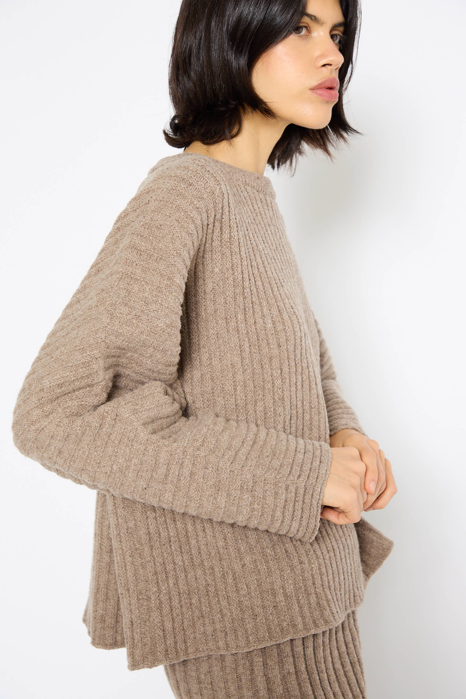 A woman with shoulder-length dark hair is wearing the Baserange Merino Wool Kai Sweater in Alassio with matching pants, standing against a white background.