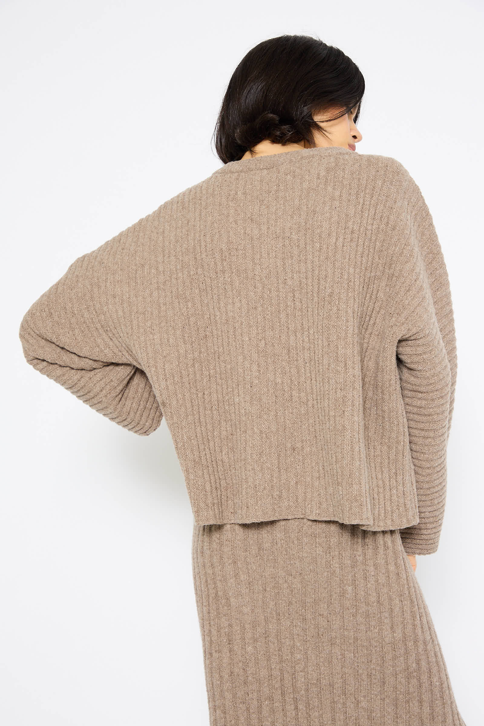 A person is shown from the back wearing a loose, ribbed Baserange Merino Wool Kai Sweater in Alassio with one arm slightly bent and their head turned to the side.