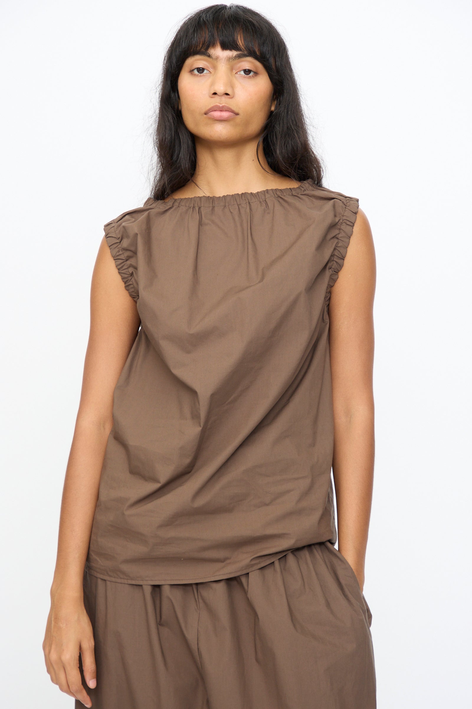 A woman faces forward against a plain white background wearing the Baserange Mia Top in Mara Brown, a sleeveless poplin top with ruffled shoulders, paired with matching loose brown pants.
