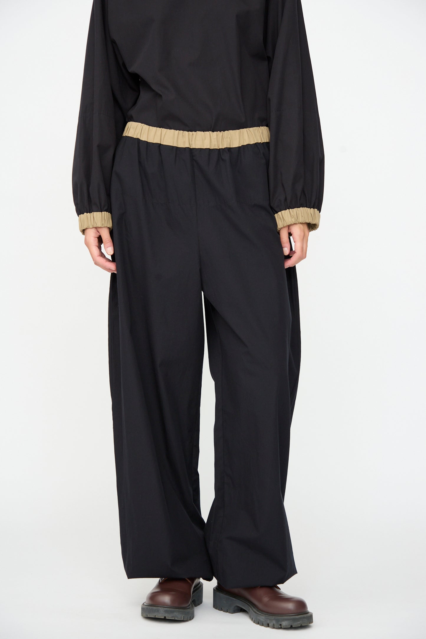 A person wearing the Baserange Moy Pant in Black Khaki—loose black cotton poplin pants with tan elastic cuffs and an elastic waistband—along with a matching top, stands on a white background. Their brown shoes are visible, but their face is not shown.