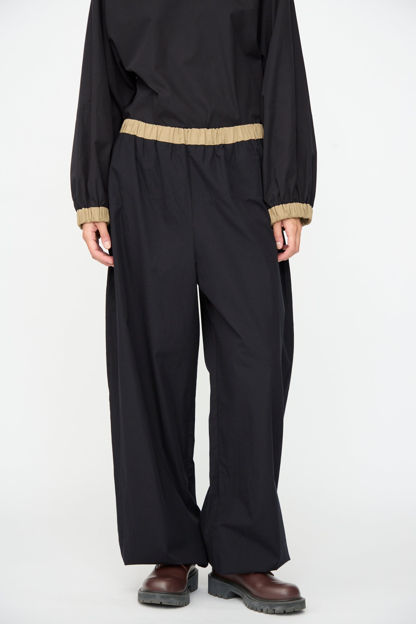 A person wearing the Baserange Moy Pant in Black Khaki—loose black cotton poplin pants with tan elastic cuffs and an elastic waistband—along with a matching top, stands on a white background. Their brown shoes are visible, but their face is not shown.