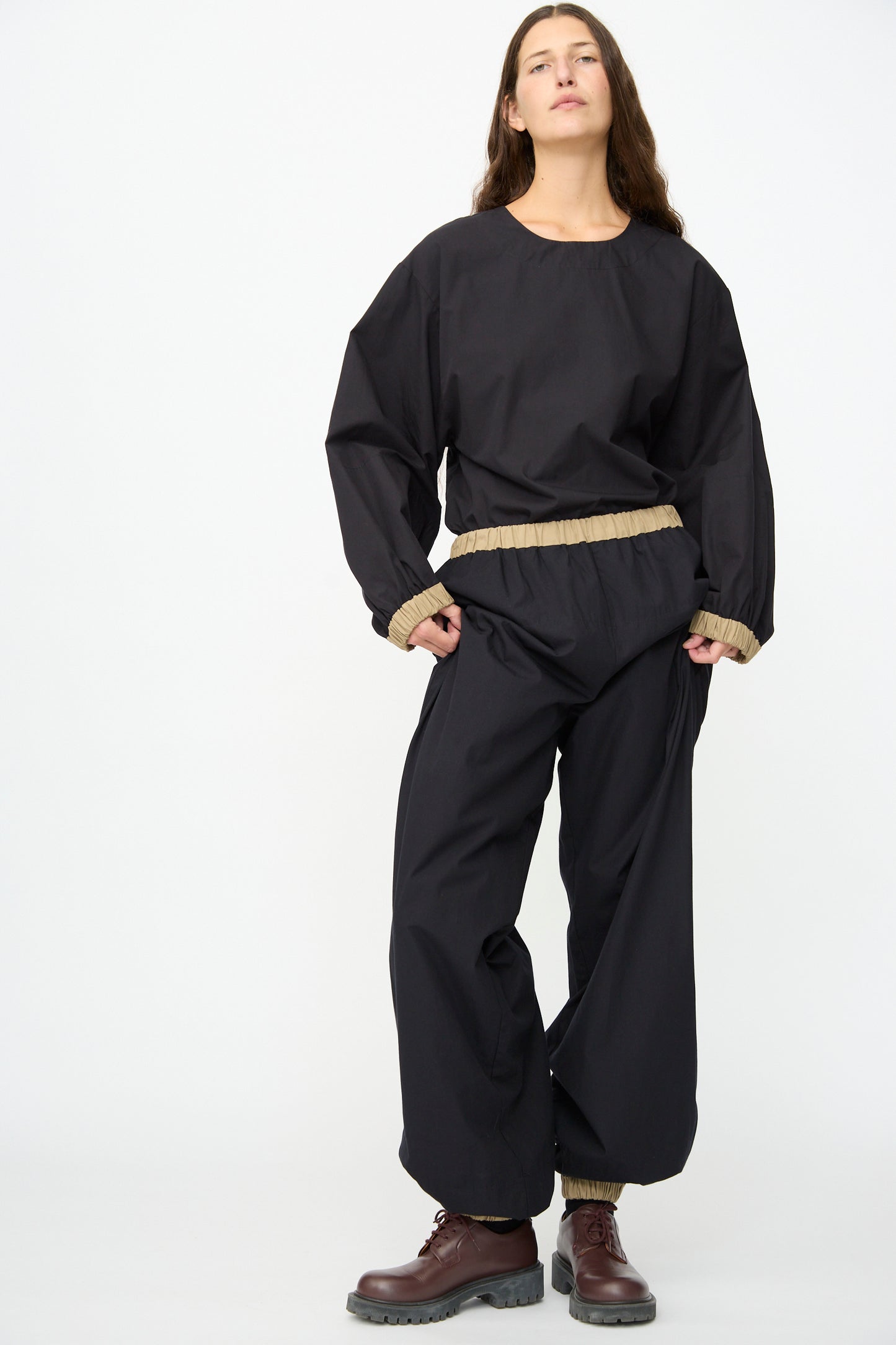 A person stands against a plain white background wearing the Baserange Moy Pant in Black Khaki paired with a loose black top and brown lace-up boots.
