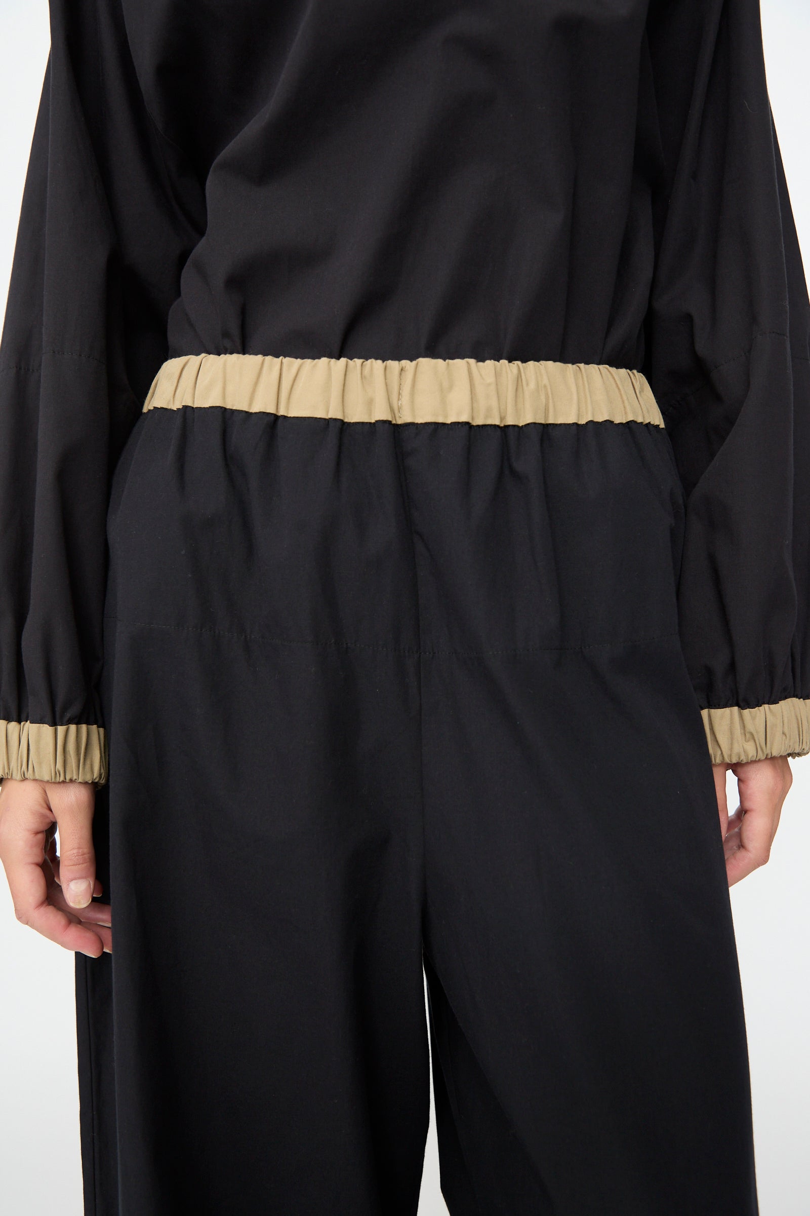 A person is shown from the back with their arms at their sides, wearing the Baserange Moy Pant in Black Khaki. The relaxed fit features tan elastic bands at the waist and cuffs, combining comfort and modern simplicity with an elastic waistband.