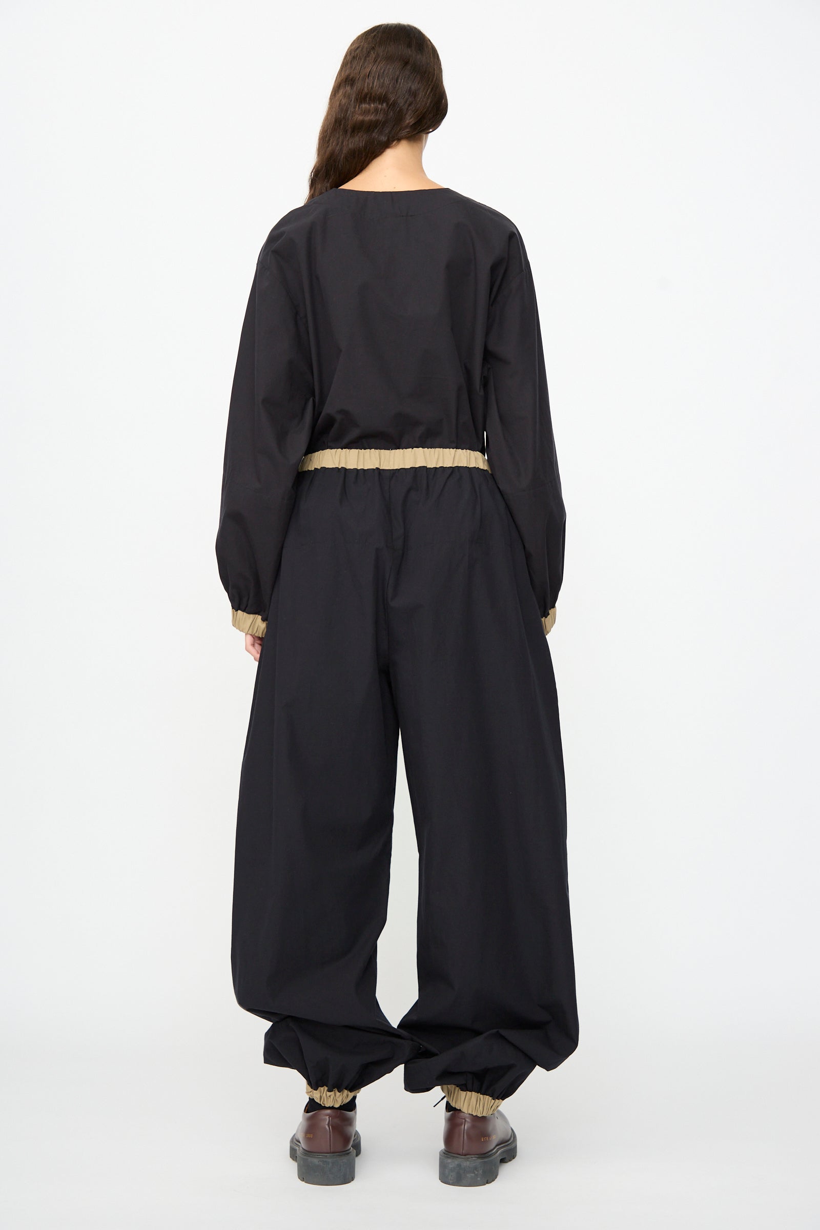 A person with long brown hair stands with their back to the camera, wearing the Baserange Moy Pant in Black Khaki—loose-fitting black cotton poplin pants featuring an elastic waistband and cuffs—paired with brown shoes.