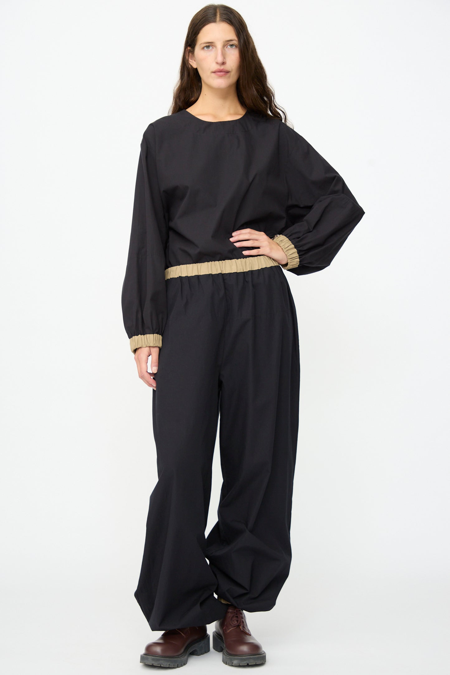 A person stands facing forward against a plain white background, wearing the Baserange Moy Pant in Black Khaki—a relaxed cotton poplin style with beige hems and an elastic waistband—paired with a loose black long-sleeve top and dark brown shoes.