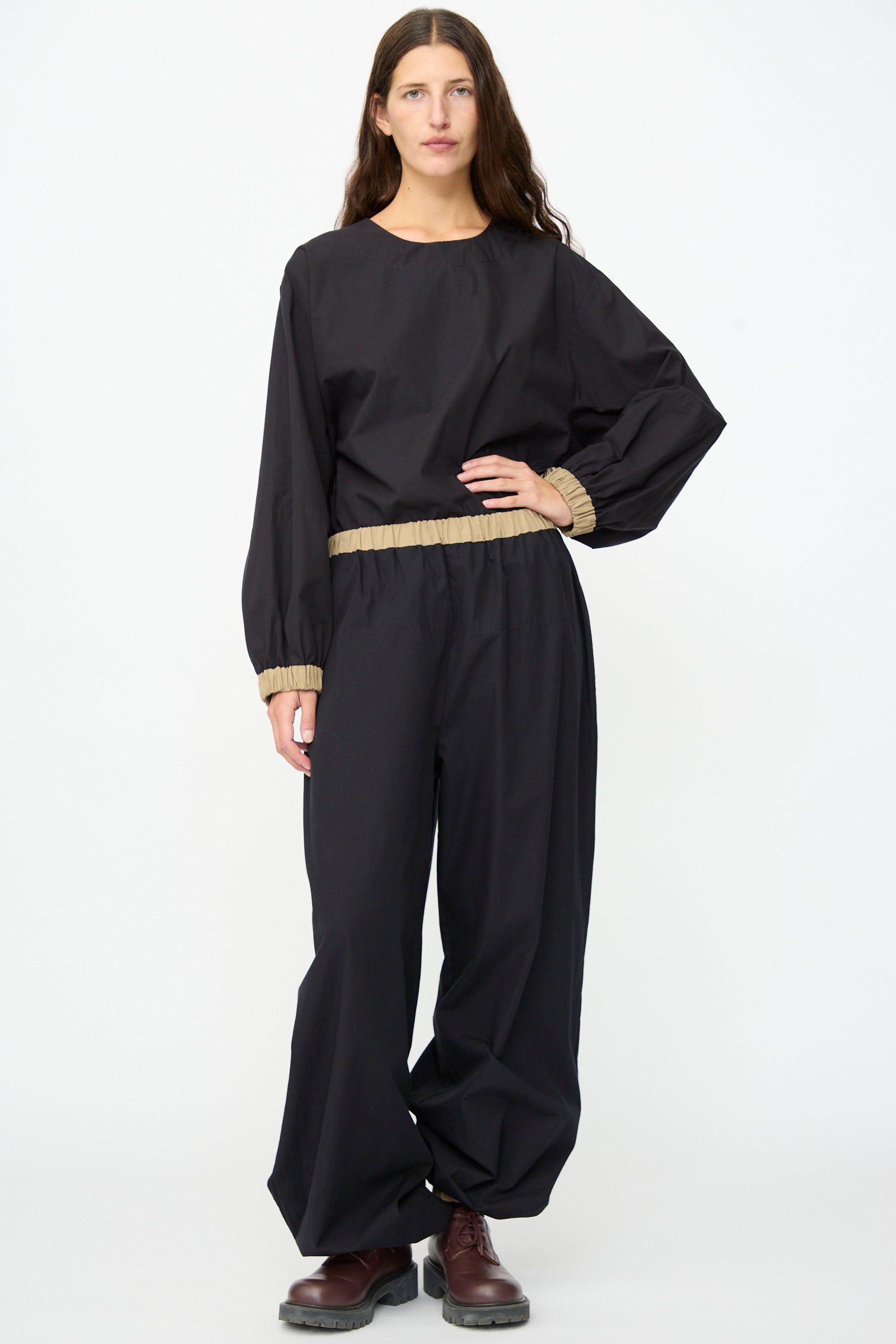A person stands facing forward against a plain white background, wearing the Baserange Moy Pant in Black Khaki—a relaxed cotton poplin style with beige hems and an elastic waistband—paired with a loose black long-sleeve top and dark brown shoes.