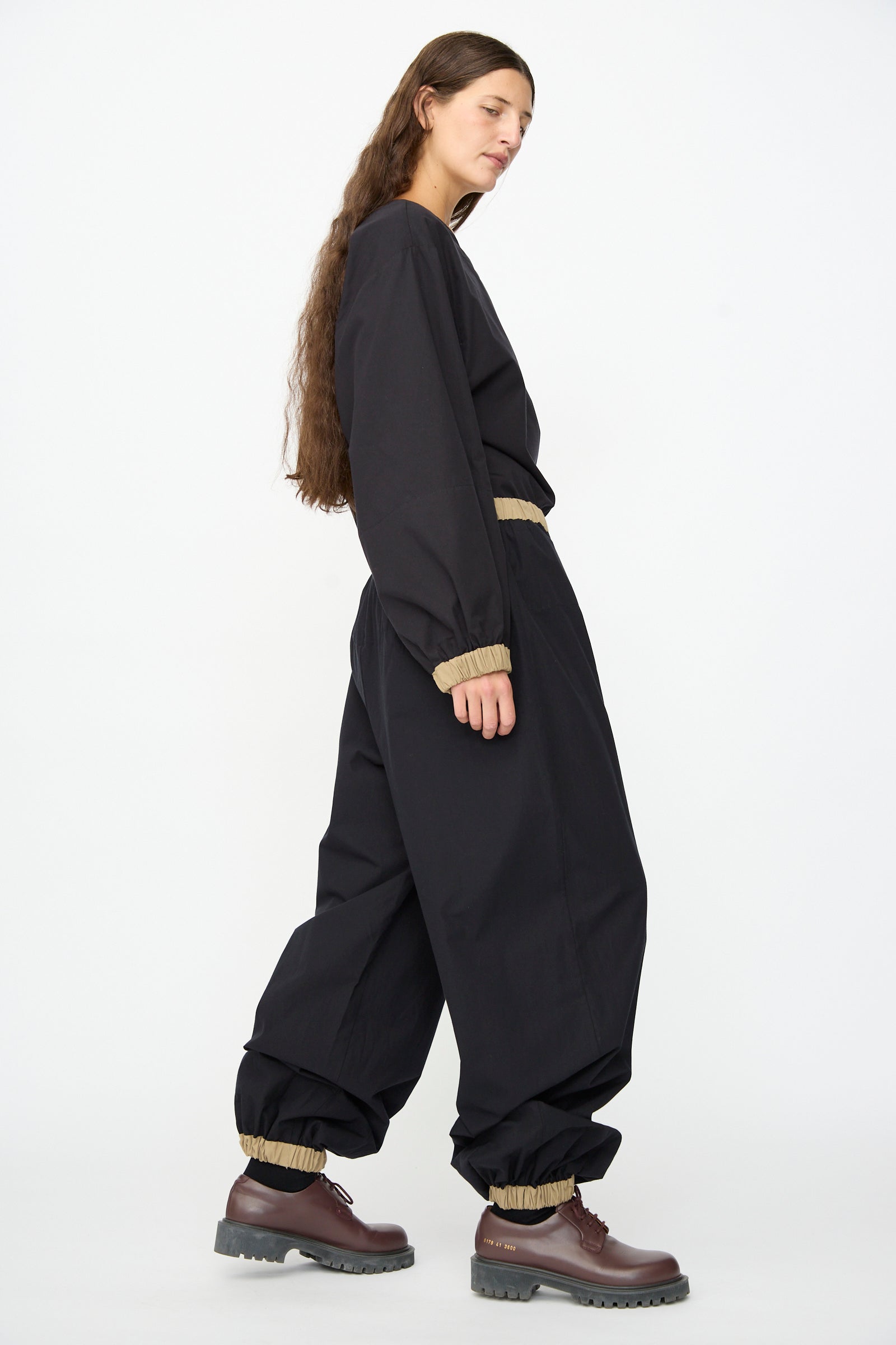 A person with long hair wears a loose black outfit featuring the Baserange Moy Pant in Black Khaki—a relaxed cotton poplin pant with a comfortable elastic waistband—paired with tan elastic cuffs at the wrists, waist, and ankles, and dark brown lace-up shoes, standing against a plain white background.