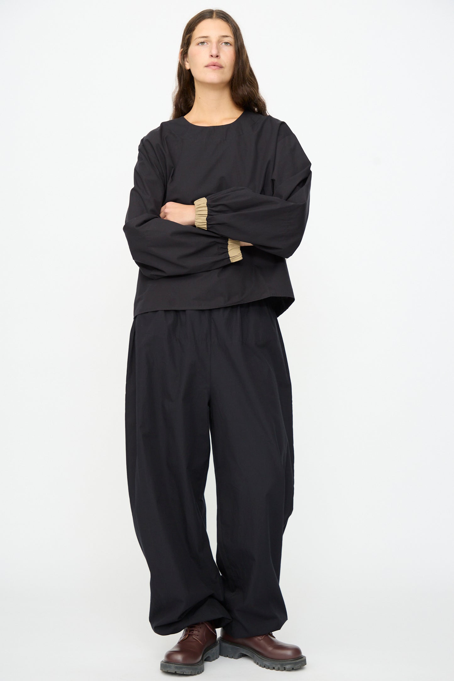 A person stands against a plain white background, wearing the Baserange Moy Top in Black Khaki with matching wide-leg pants and brown shoes, arms crossed and a neutral expression.