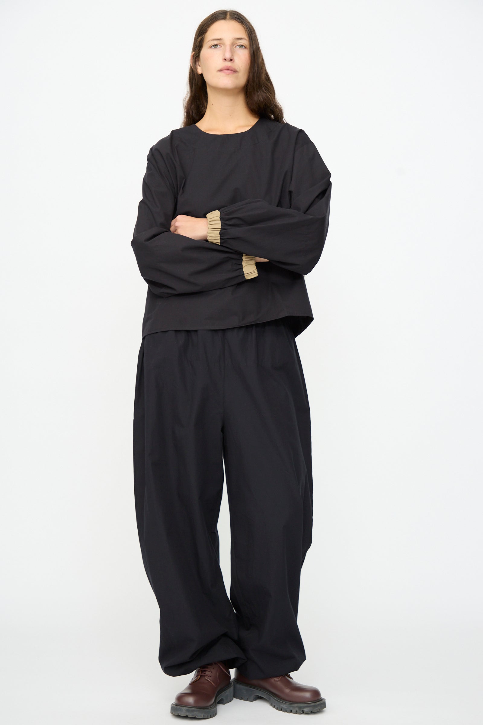 A person stands against a plain white background, wearing the Baserange Moy Top in Black Khaki with matching wide-leg pants and brown shoes, arms crossed and a neutral expression.