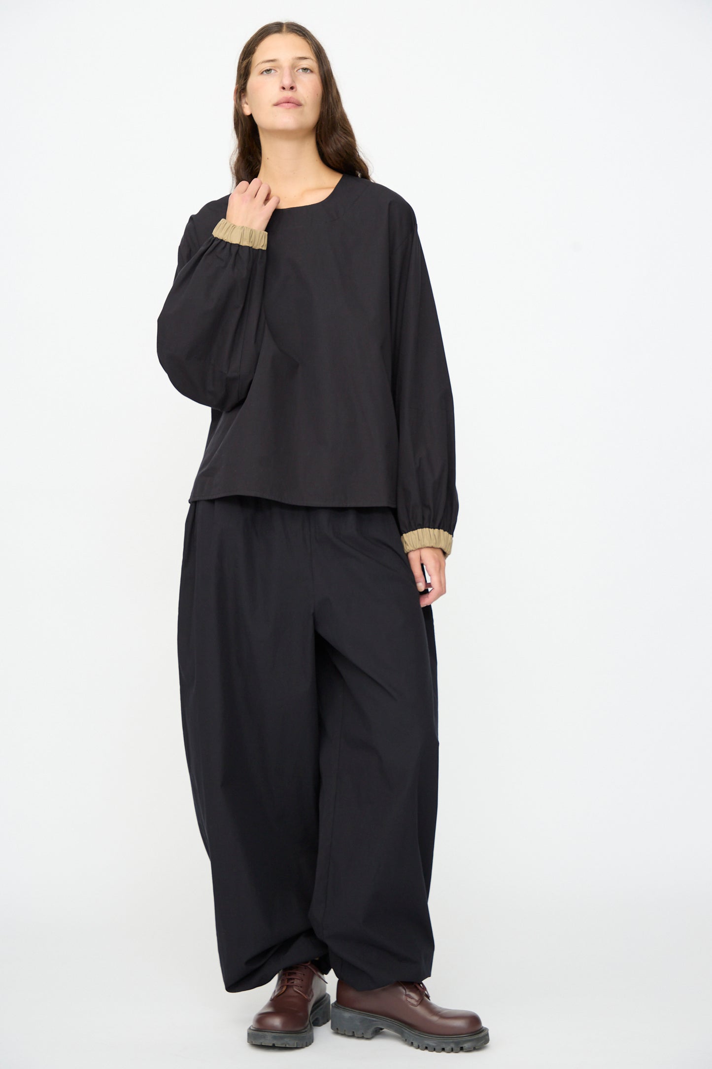 A person stands in front of a white background, wearing the Baserange Moy Top in Black Khaki—a relaxed fit, long sleeve cotton poplin top—paired with wide black pants, brown shoes, and a neutral expression.