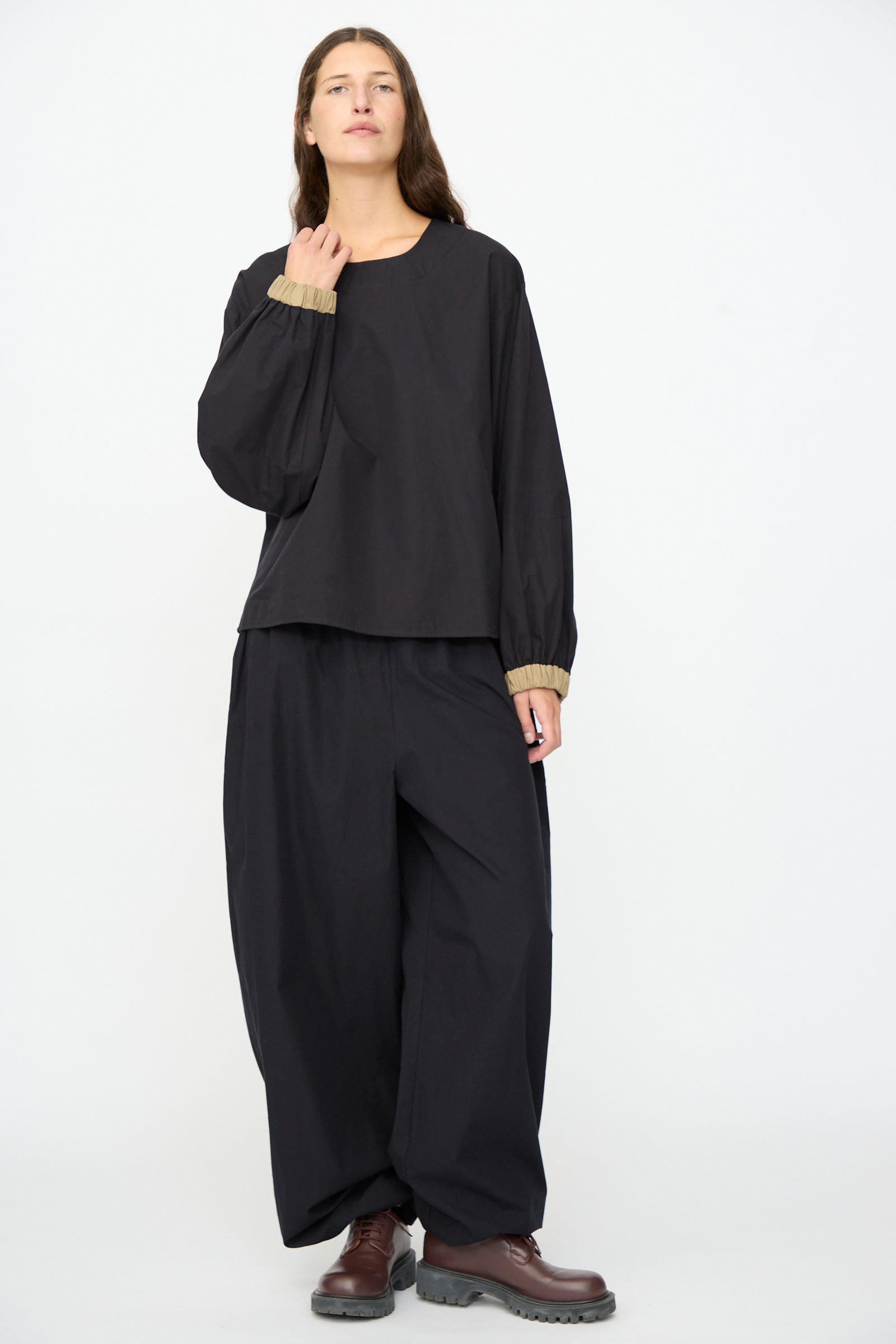 A person stands in front of a white background, wearing the Baserange Moy Top in Black Khaki—a relaxed fit, long sleeve cotton poplin top—paired with wide black pants, brown shoes, and a neutral expression.