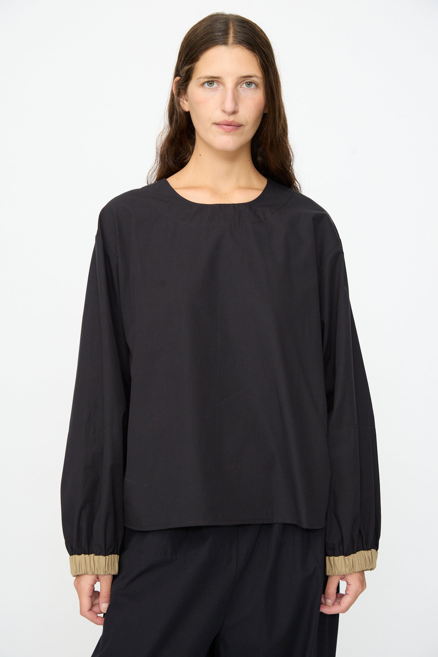 A person with long brown hair wears the Baserange Moy Top in Black Khaki, a relaxed fit black long sleeve top with beige elastic cuffs, paired with matching black pants, standing against a plain white background.