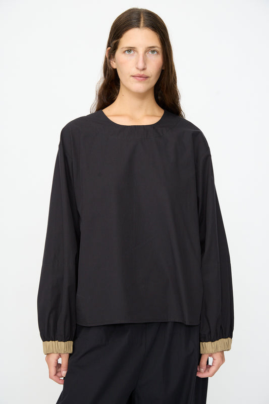 A person with long brown hair wears the Baserange Moy Top in Black Khaki, a relaxed fit black long sleeve top with beige elastic cuffs, paired with matching black pants, standing against a plain white background.