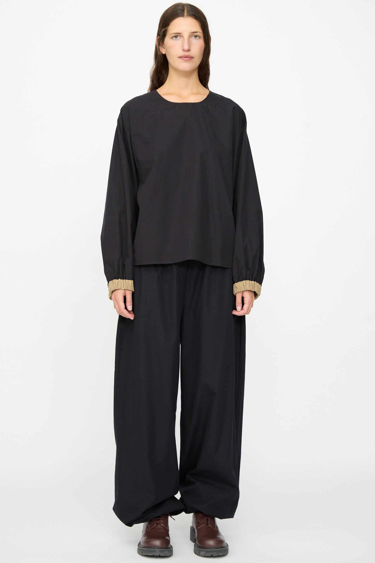 A person faces forward against a plain white background, wearing the Baserange Moy Top in Black Khaki with matching wide-leg pants and brown shoes.