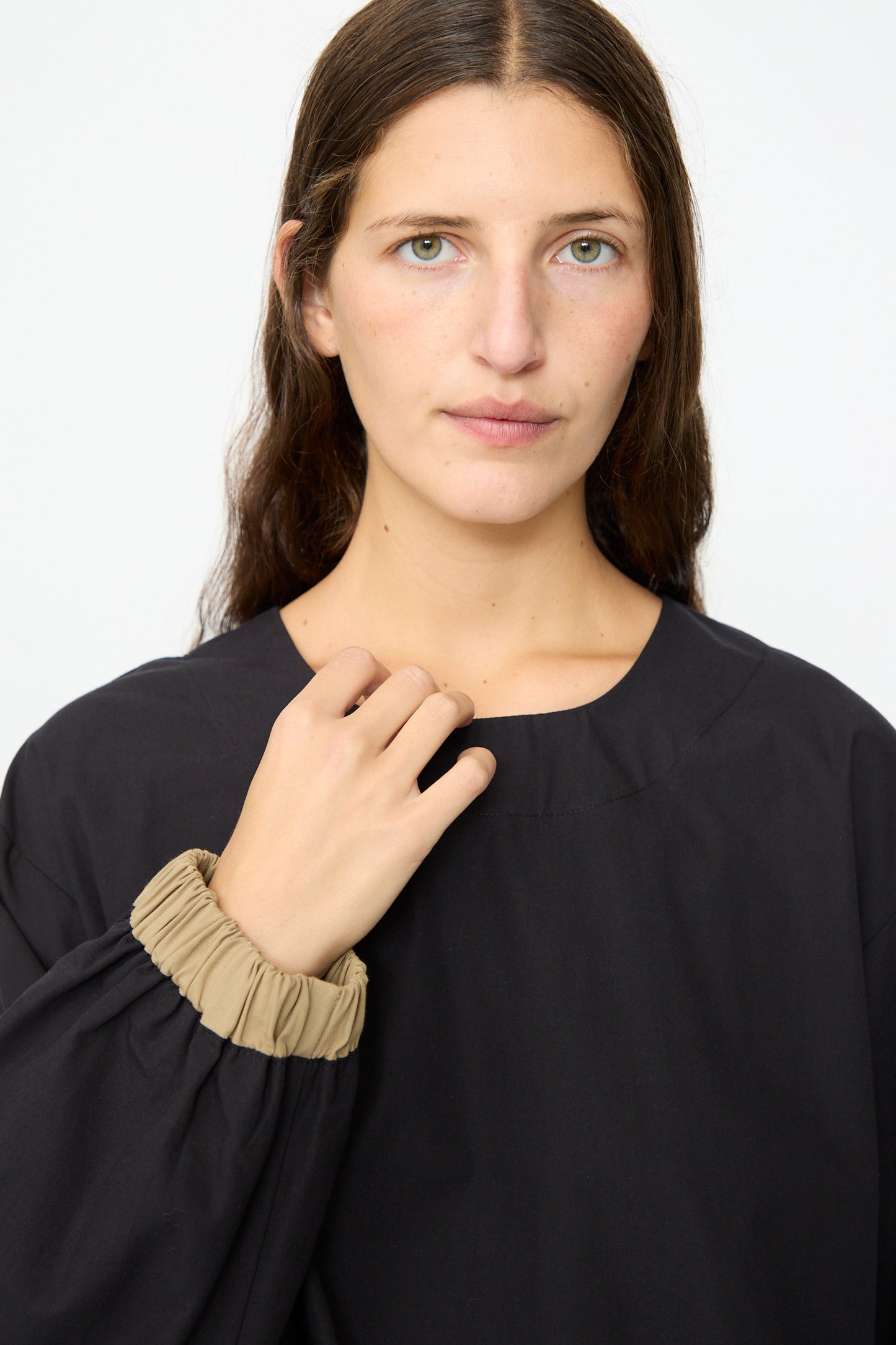 A person with long brown hair wears the Baserange Moy Top in Black Khaki—a black cotton poplin top with beige elastic cuffs and a relaxed fit—as they look at the camera against a plain white background.