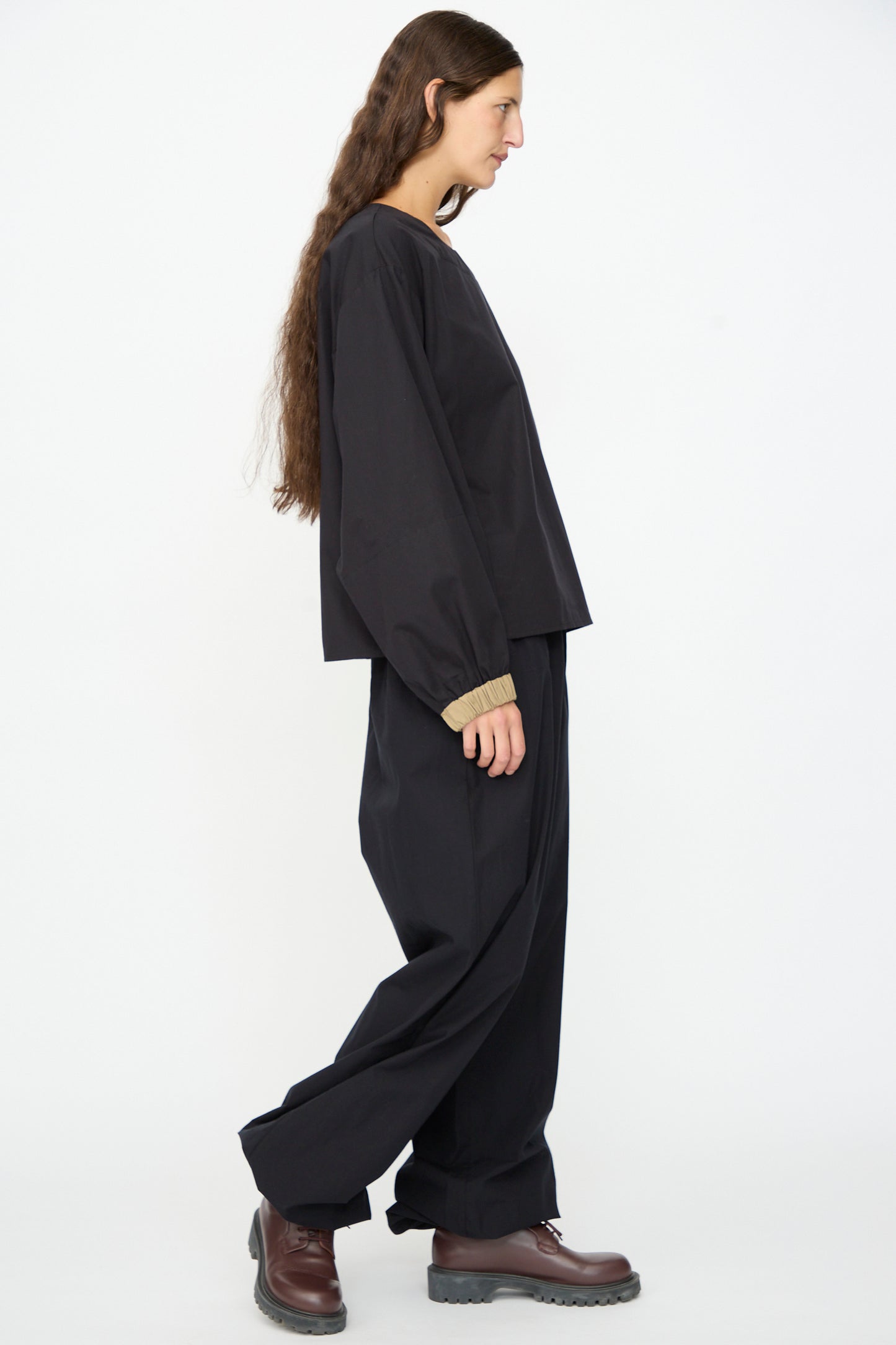 A person with long brown hair, wearing the Baserange Moy Top in Black Khaki and matching relaxed-fit pants with brown boots, stands in profile against a white background.