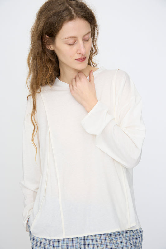 Wearing the Baserange Mul Long Sleeve Tee in Off White, a person with long wavy hair and blue plaid pants stands against a plain light background, looking down with one hand near their neck.