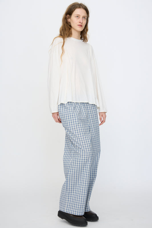 A person stands against a plain white background wearing the Baserange Mul Long Sleeve Tee in Off White, paired with blue and white plaid wide-leg pants and black shoes.