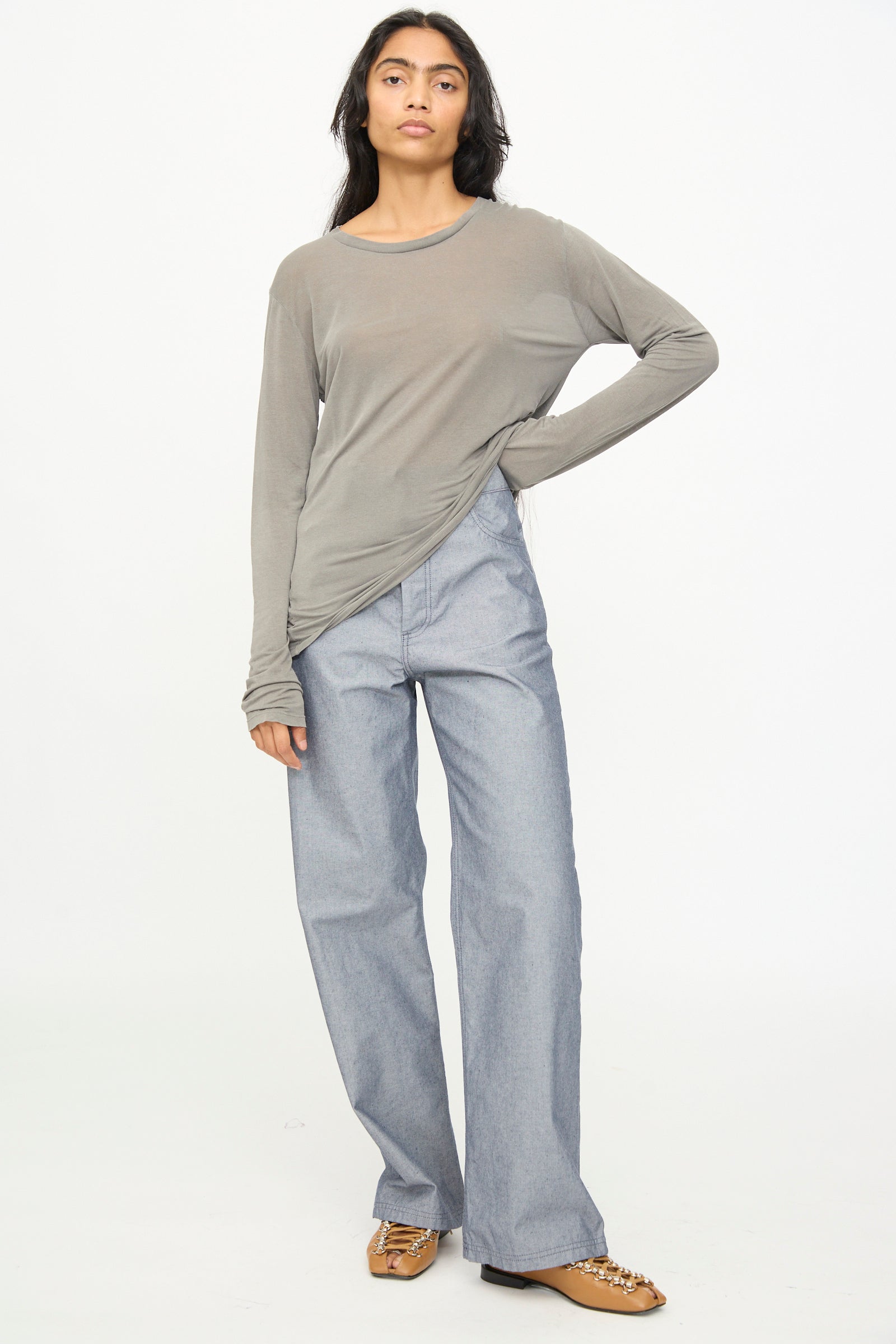 A person stands facing forward against a plain white background, wearing a loose grey long-sleeve shirt, the Baserange Navalo Pants in Dark Denim—high-rise pants crafted from recycled denim—and brown shoes with chain details.