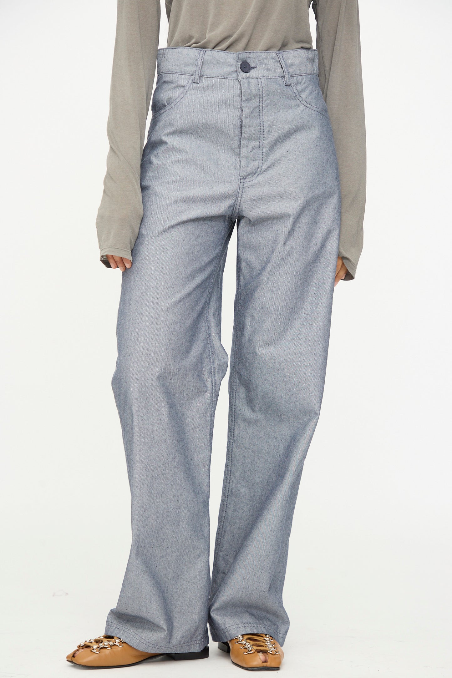 A person wearing a gray long-sleeve shirt, Baserange Navalo Pants in Dark Denim crafted from recycled denim, and tan flat shoes with gold chain detail stands against a plain white background.
