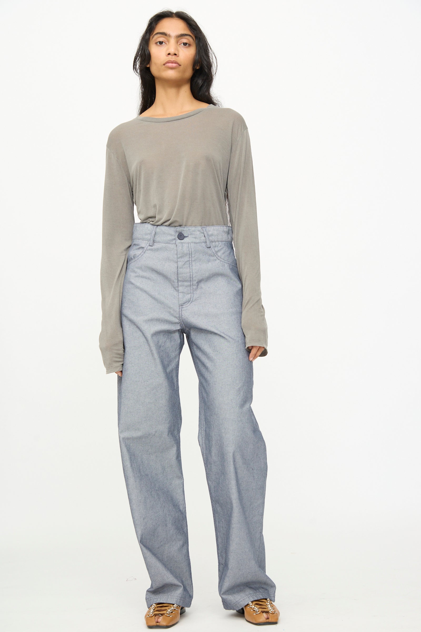 A person stands facing forward against a plain white background, wearing a loose grey long-sleeve shirt tucked into Baserange Navalo Pants in Dark Denim—high-rise, wide-leg pants made from recycled denim—and brown shoes.
