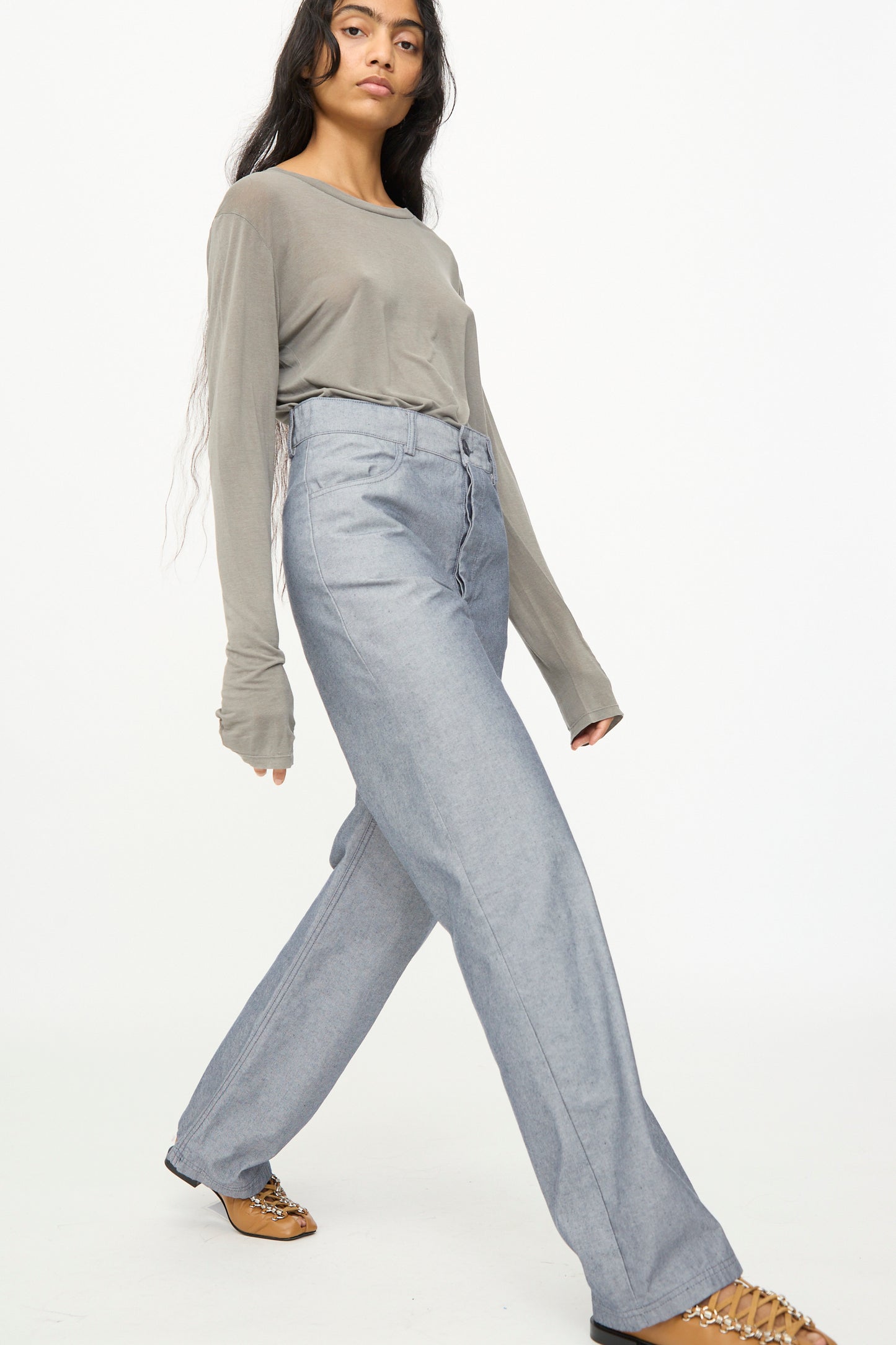 A person wearing a loose long-sleeve gray organic cotton shirt, paired with the Baserange Navalo Pants in Dark Denim—a high-waisted, wide-leg style crafted from recycled denim—and brown heeled shoes, walking against a plain white background.