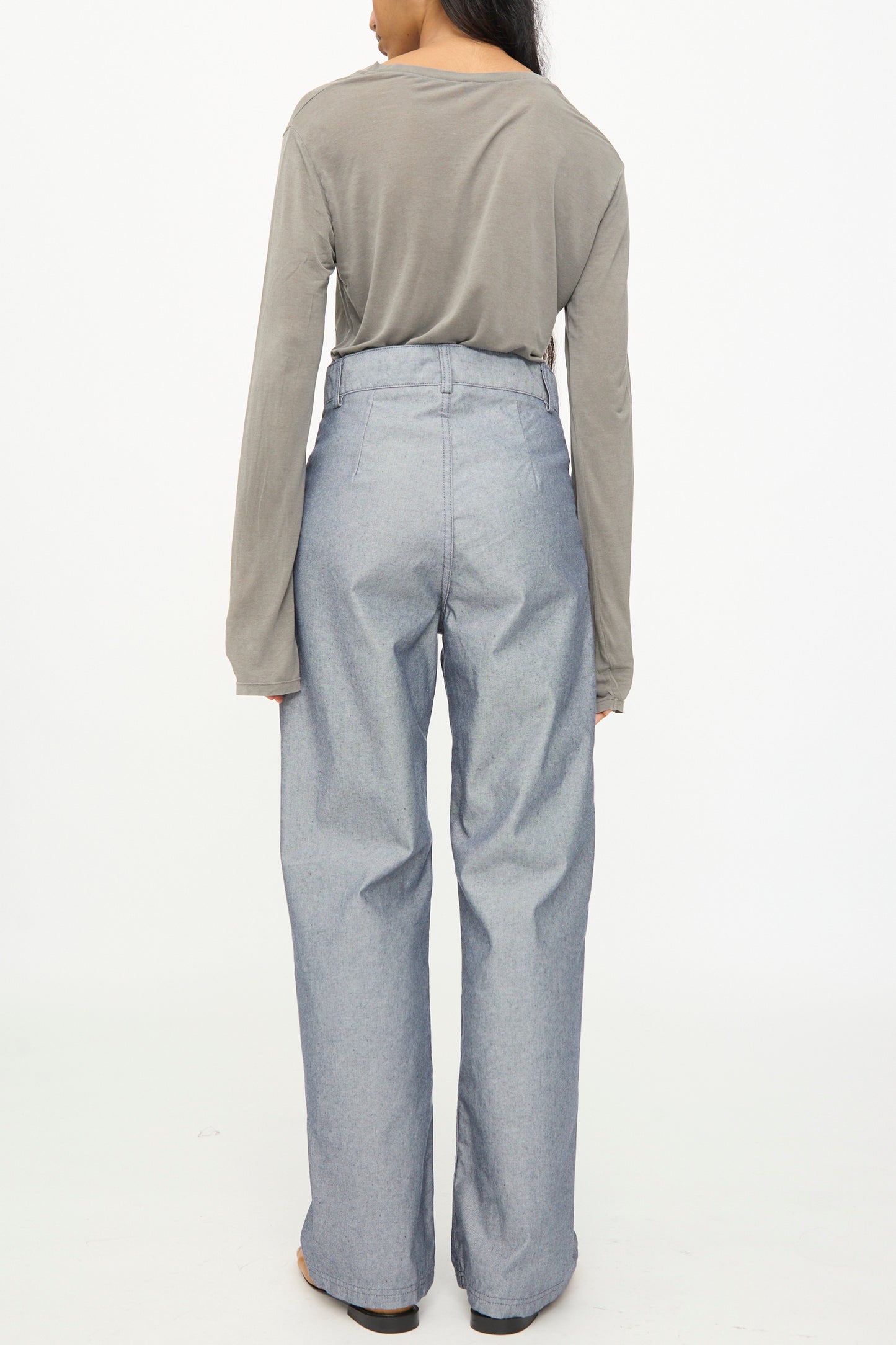 A person standing facing away, wearing a long-sleeved gray shirt and the Baserange Navalo Pants in Dark Denim—high rise, loose-fitting pants made from recycled denim—against a plain white background.