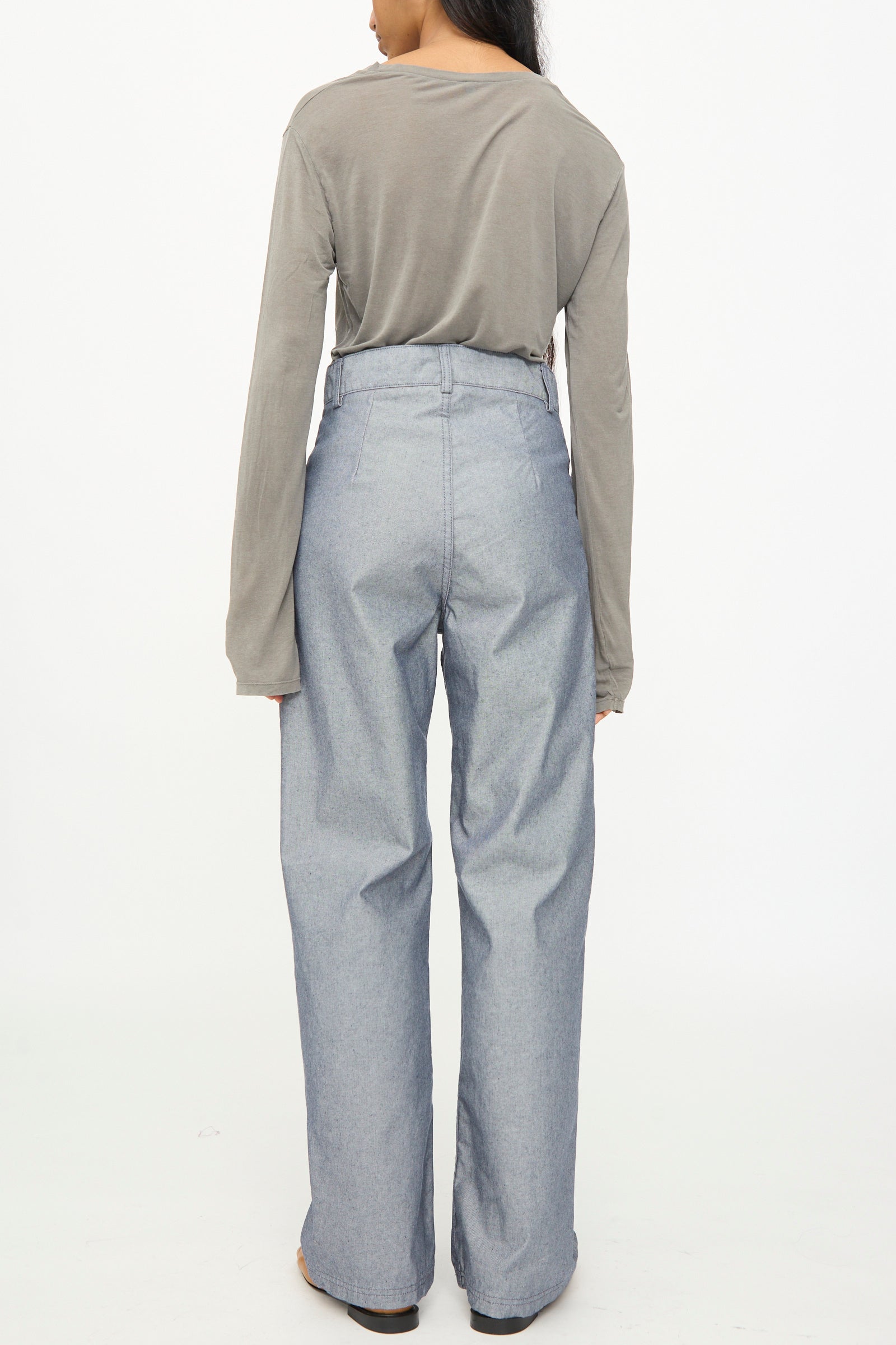 A person standing facing away, wearing a long-sleeved gray shirt and the Baserange Navalo Pants in Dark Denim—high rise, loose-fitting pants made from recycled denim—against a plain white background.