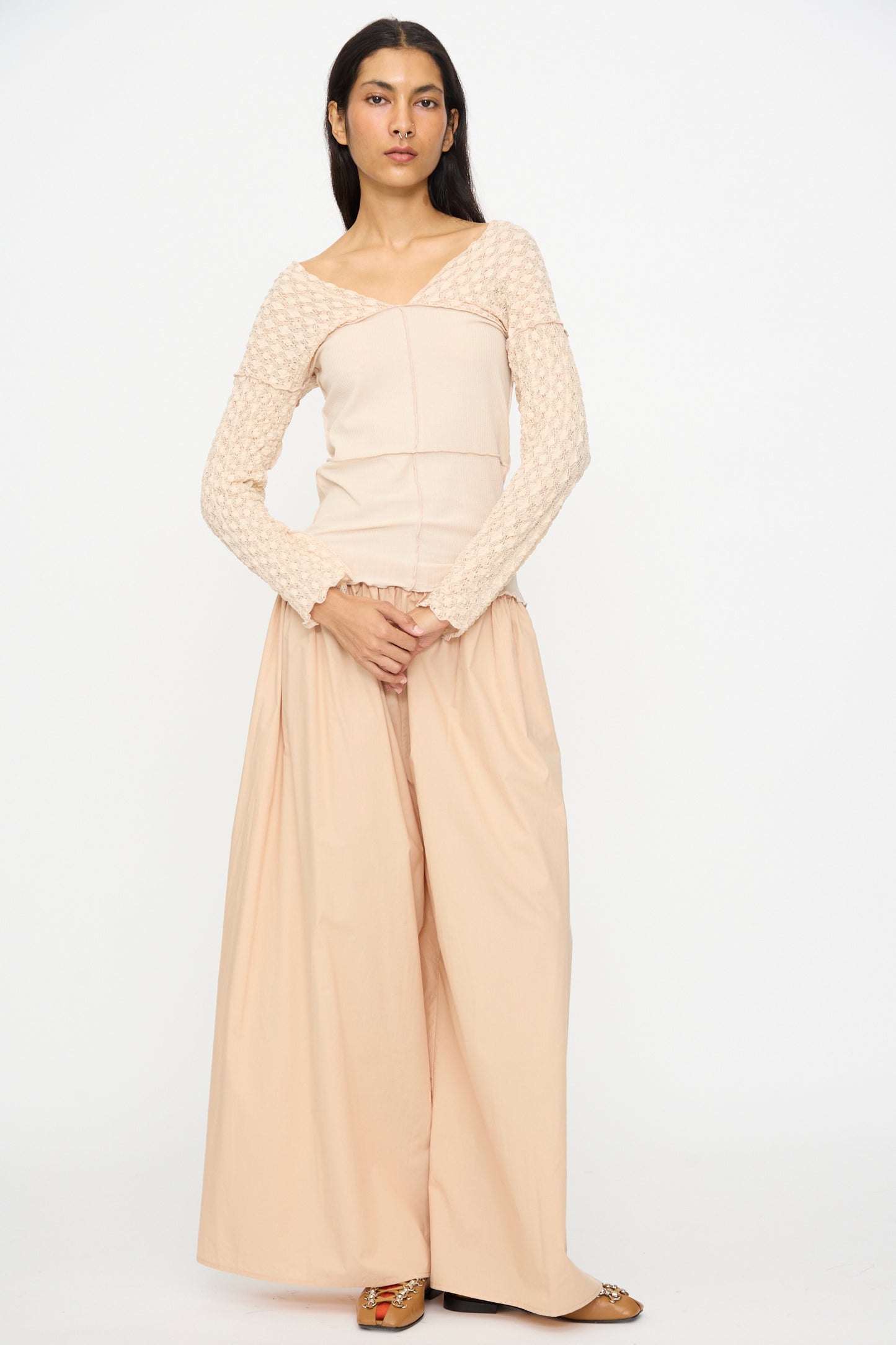 A woman stands against a white background wearing the Baserange Nehru Long Sleeve Tee in Dylan Beige, paired with wide-leg beige pants and flat shoes.