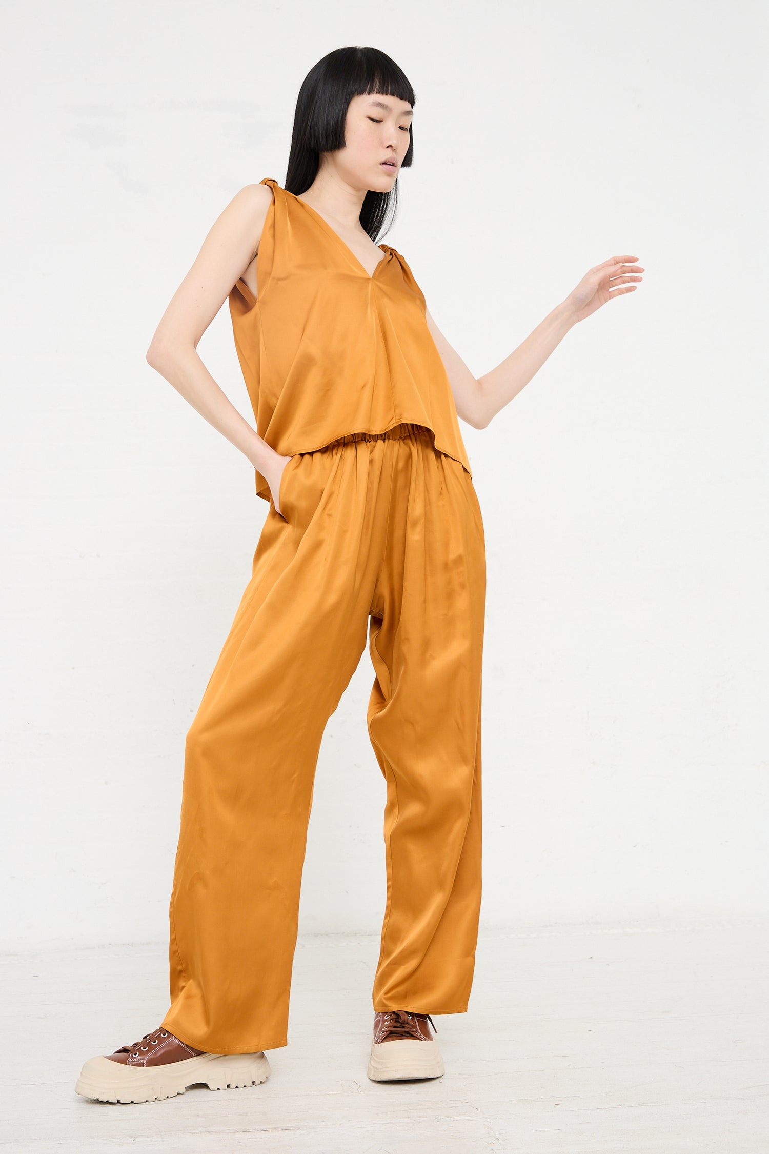 A person in a Neil Top in Mina Copper by Baserange and brown shoes posing with one hand raised against a plain backdrop.