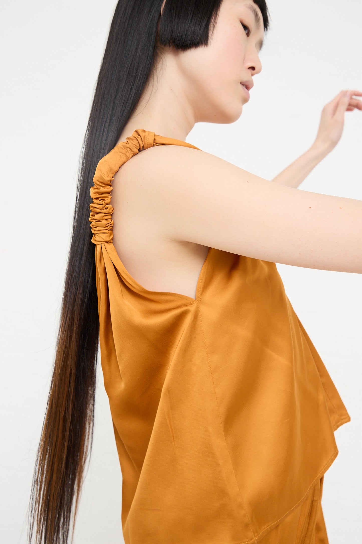 Woman wearing an orange Neil Top in Mina Copper by Baserange, a V-neck tank top with ruched shoulder detail, viewed from the side.