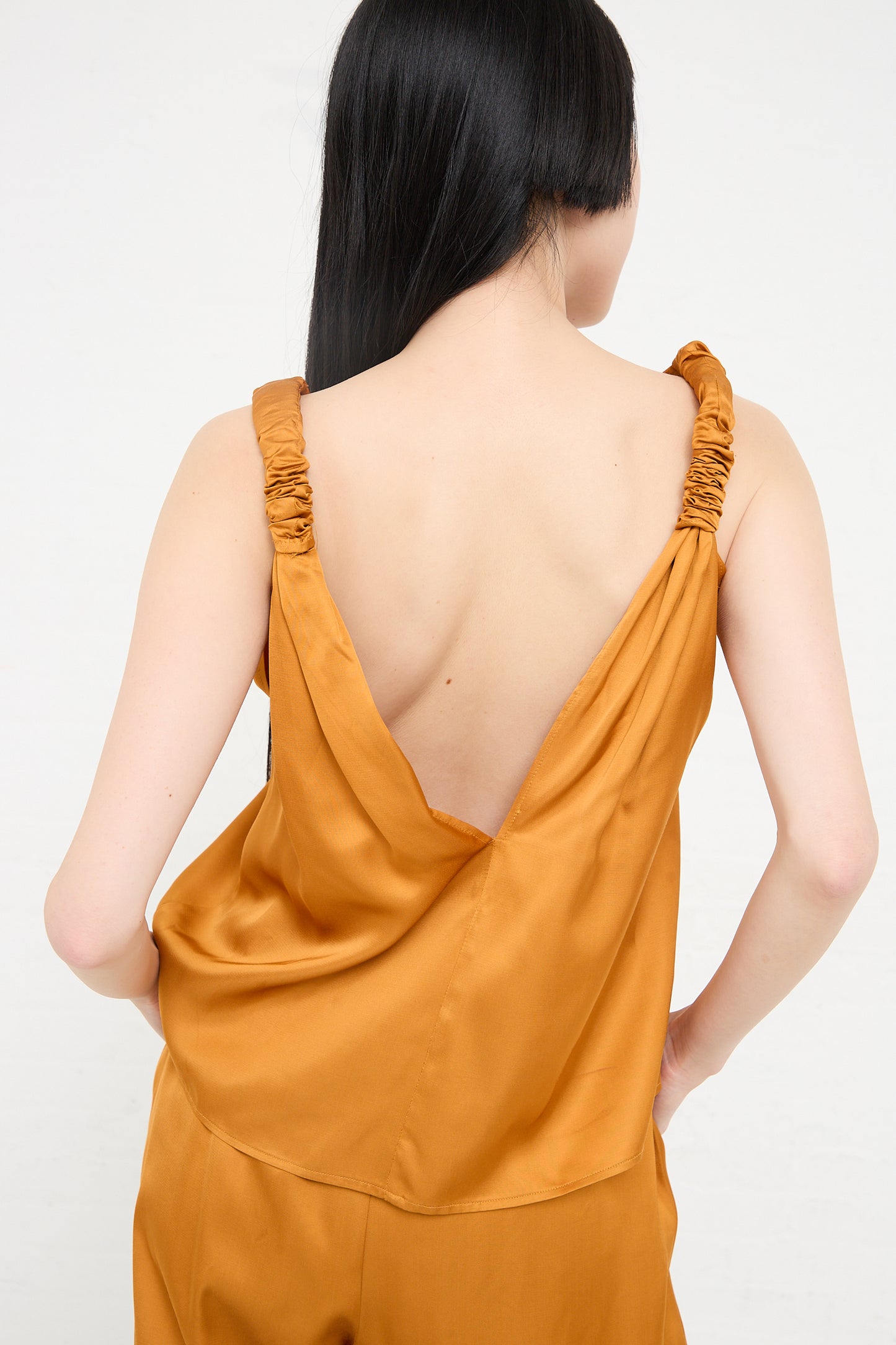 Woman wearing an orange Neil Top in Mina Copper by Baserange, a V-neck tank top with ruched shoulder detail,  back view.