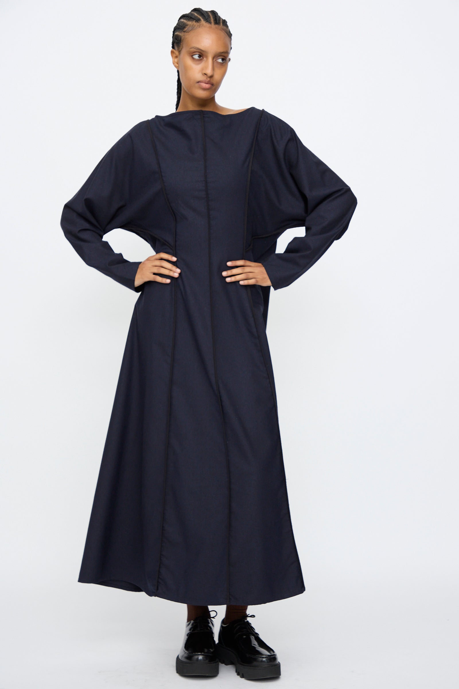 A person stands against a white background wearing the Baserange Nika Dress in Dama Navy, a long-sleeve maxi dress with a boat neck, paired with black platform shoes.