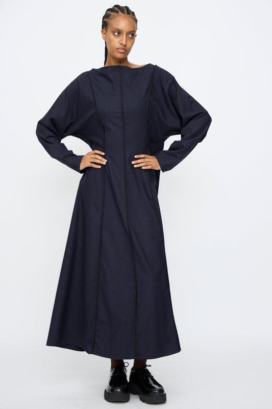 A person stands against a white background wearing the Baserange Nika Dress in Dama Navy, a long-sleeve maxi dress with a boat neck, paired with black platform shoes.