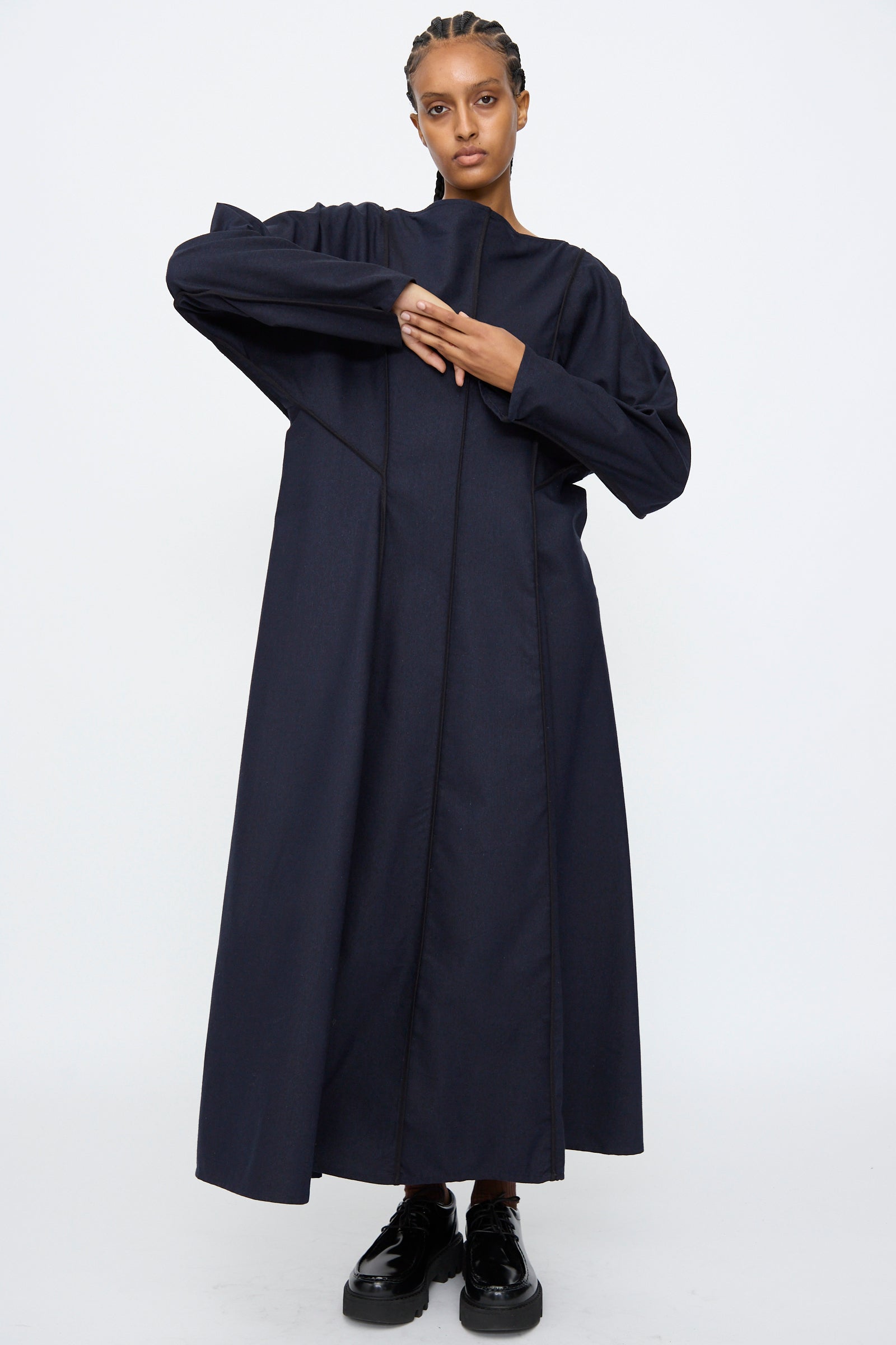 A person stands against a plain background wearing the Baserange Nika Dress in Dama Navy, a long sleeve silk wool blend maxi with a boat neck, paired with black shoes and arms positioned asymmetrically.