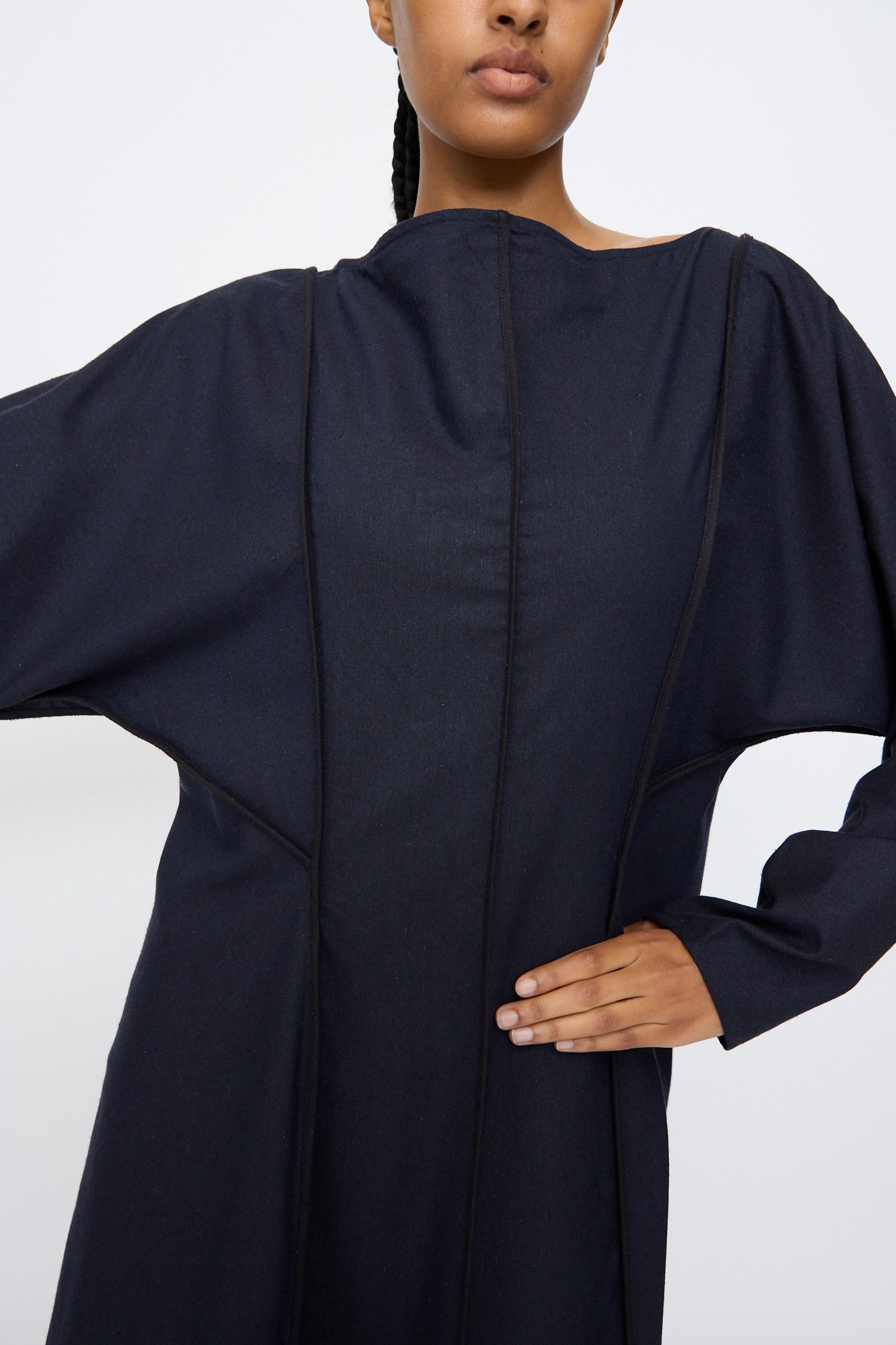 A person in the Baserange Nika Dress in Dama Navy, a long sleeve maxi with seam lines and a boat neck, stands with one hand on their hip against a plain background.