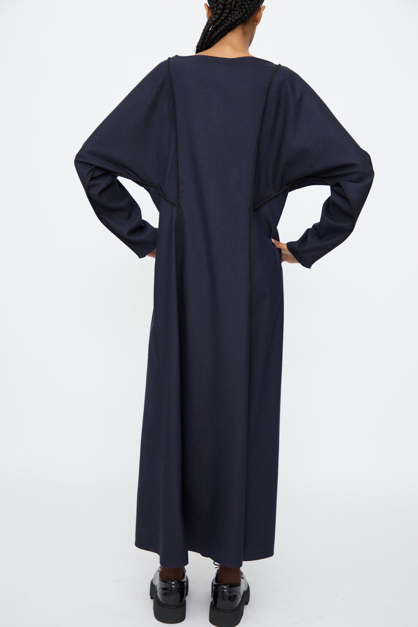 A person stands with their back to the camera, wearing the Baserange Nika Dress in Dama Navy, a long sleeve maxi dress, paired with black shoes against a plain white background.
