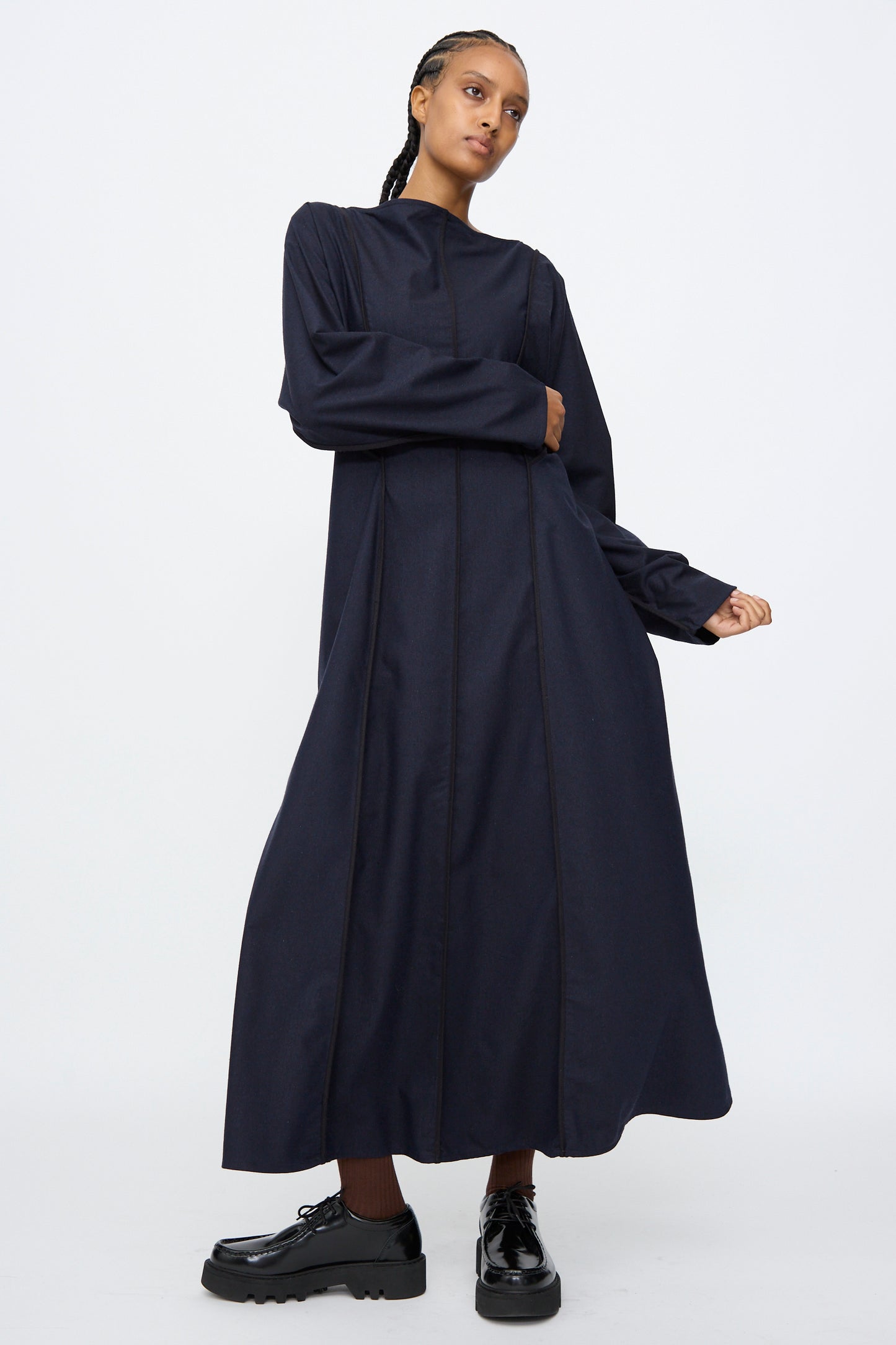 A person wearing the Baserange Nika Dress in Dama Navy—a navy, long sleeve, silk wool blend maxi dress with a boat neck—and black platform shoes stands against a plain white background.